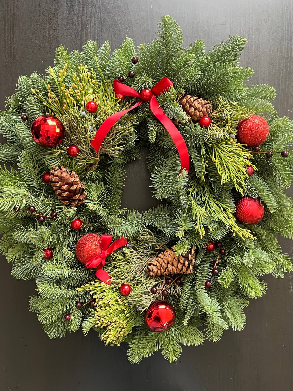 Christmas Charm Christmas Charm - Christmas Charm is the embodiment of warmth, tradition, and holiday magic. This elegant Christmas wreath is crafted from fresh evergreens, accented with natural pinecones and adorned with vibrant red ornaments and berries. A graceful crimson bow adds a touch of sophistication, creating an atmosphere of joy and festivity. Christmas Charm is the perfect decoration for your door, fireplace, or holiday table, bringing a sense of winter wonder and Christmas coziness into your home.