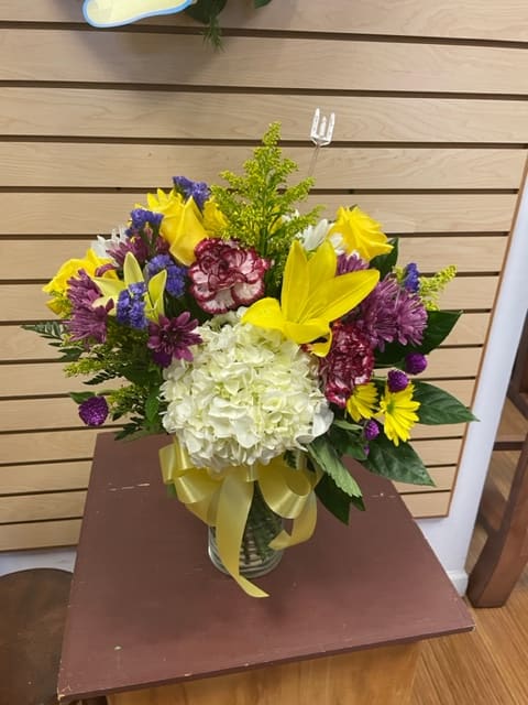 Bright Wishes Bright Wishes - Mixed cut arrangement with yellow lilies, yellow roses, purple daisies, white hydrangea, purple statice, white daisies, and bicolor carnations