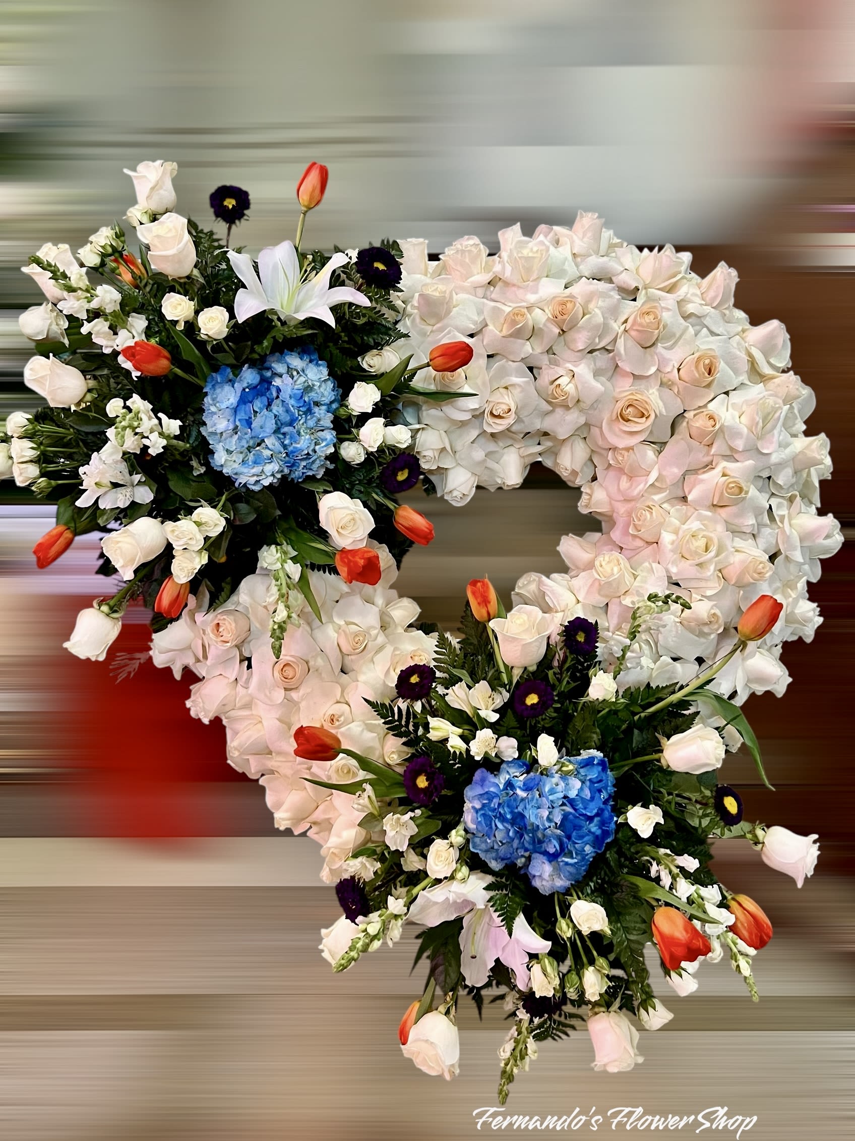 Peaceful Passage - This unique floral arrangement is a beautifully crafted heart design, symbolizing love and remembrance. The base features soft cream and blush roses, creating a classic and elegant foundation. Vibrant pops of color are introduced with bright blue hydrangeas, deep purple asters, and striking orange tulips, adding a lively yet balanced contrast. Delicate white lilies and lush greenery enhance the overall composition, bringing texture and depth. Displayed on a rustic wooden backdrop, this design from “Fernando’s Flower Shop” is perfect for expressing heartfelt emotions and honoring cherished memories at special events or memorial services.