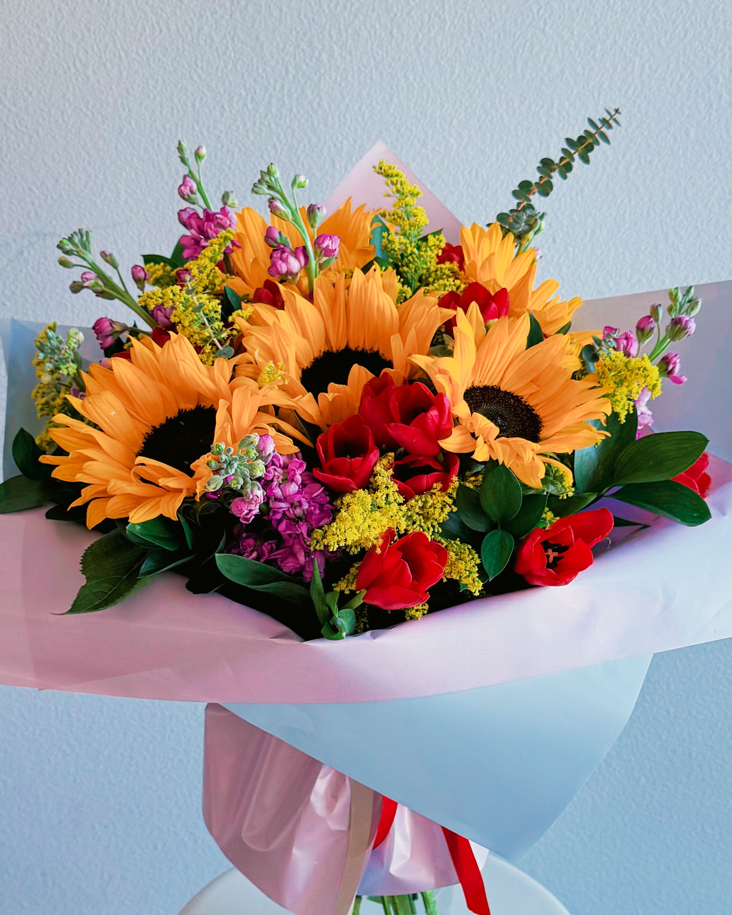 Bright Bloom - This bouquet is like a burst of sunshine wrapped in love. With bright yellow sunflowers, deep red tulips, and pops of purple and green, it’s got that perfect mix of warmth and vibrancy. It’s giving pure joy, summer vibes, and a whole lotta romance. The mix of textures and colors makes it look fresh, lively, and effortlessly chic—kinda like that perfect outfit that turns heads without even trying.