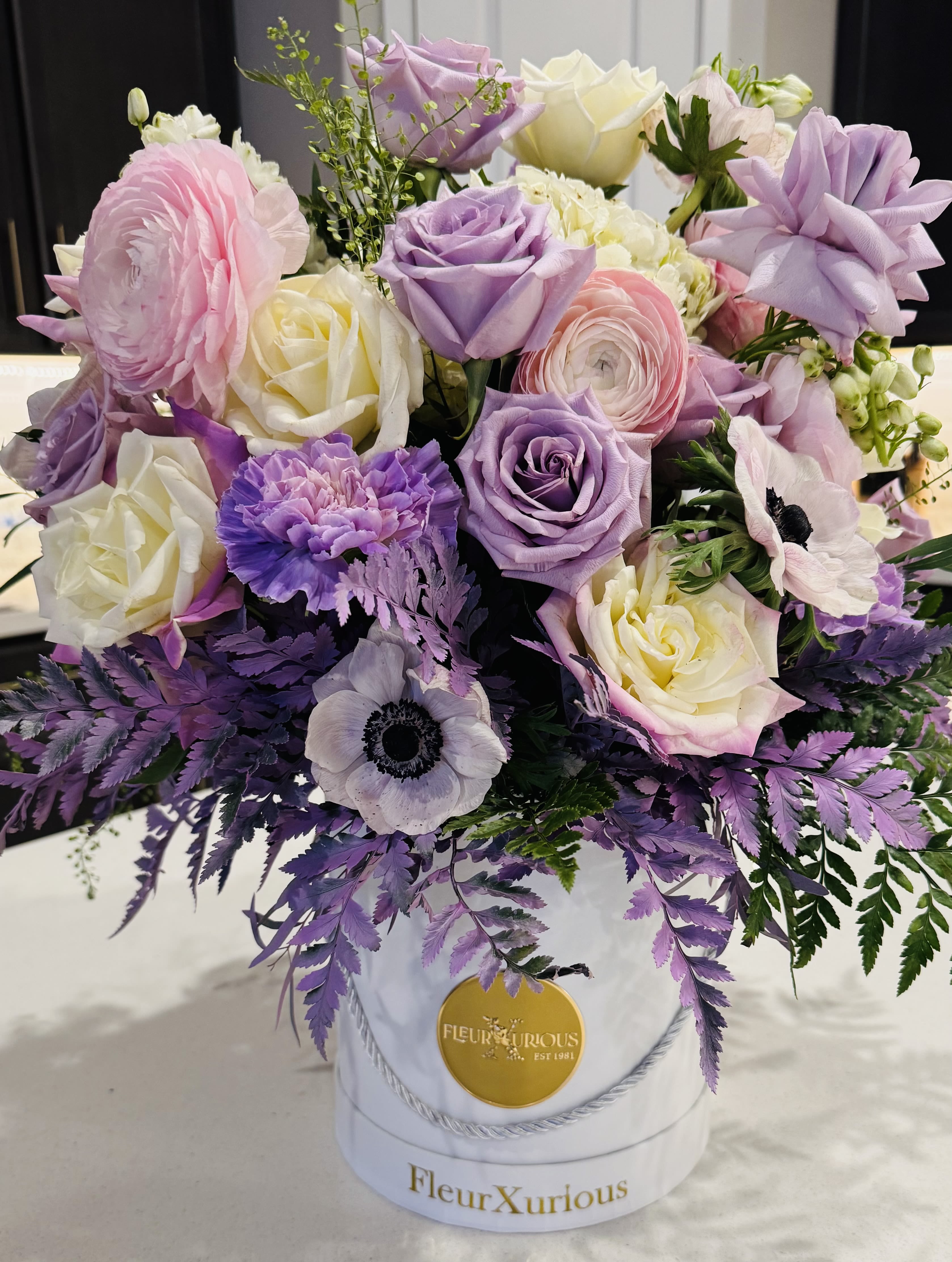 A Purple Passion - Anemones, Rannunculas, Light purple roses, white hydrangeas, purple Carnations, with a touch of greens lightly sprayed purple leather leaf tucked in customer logo FleurXurious box. (Some flowers in this arrangement are seasonal. Check for availability)