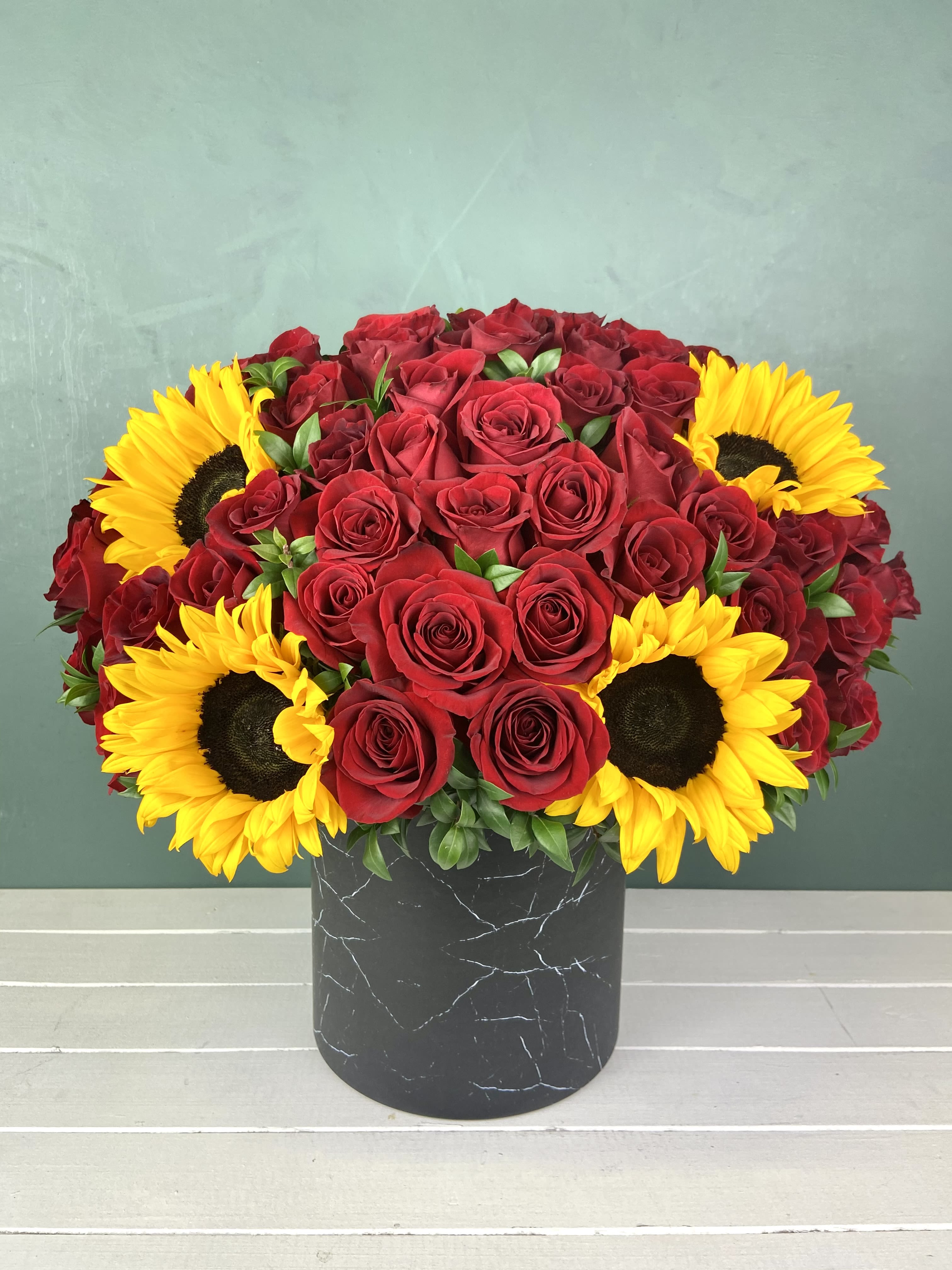 82D- Sunset Romance - This striking arrangement showcases bold red roses paired with vibrant yellow sunflowers. The contrast between the rich red and bright yellow creates an eye-catching, cheerful display. Set in a modern, marbled black vase, this design blends elegance with warmth, making it perfect for special celebrations or to brighten any space. DELUXE size is shown in the picture