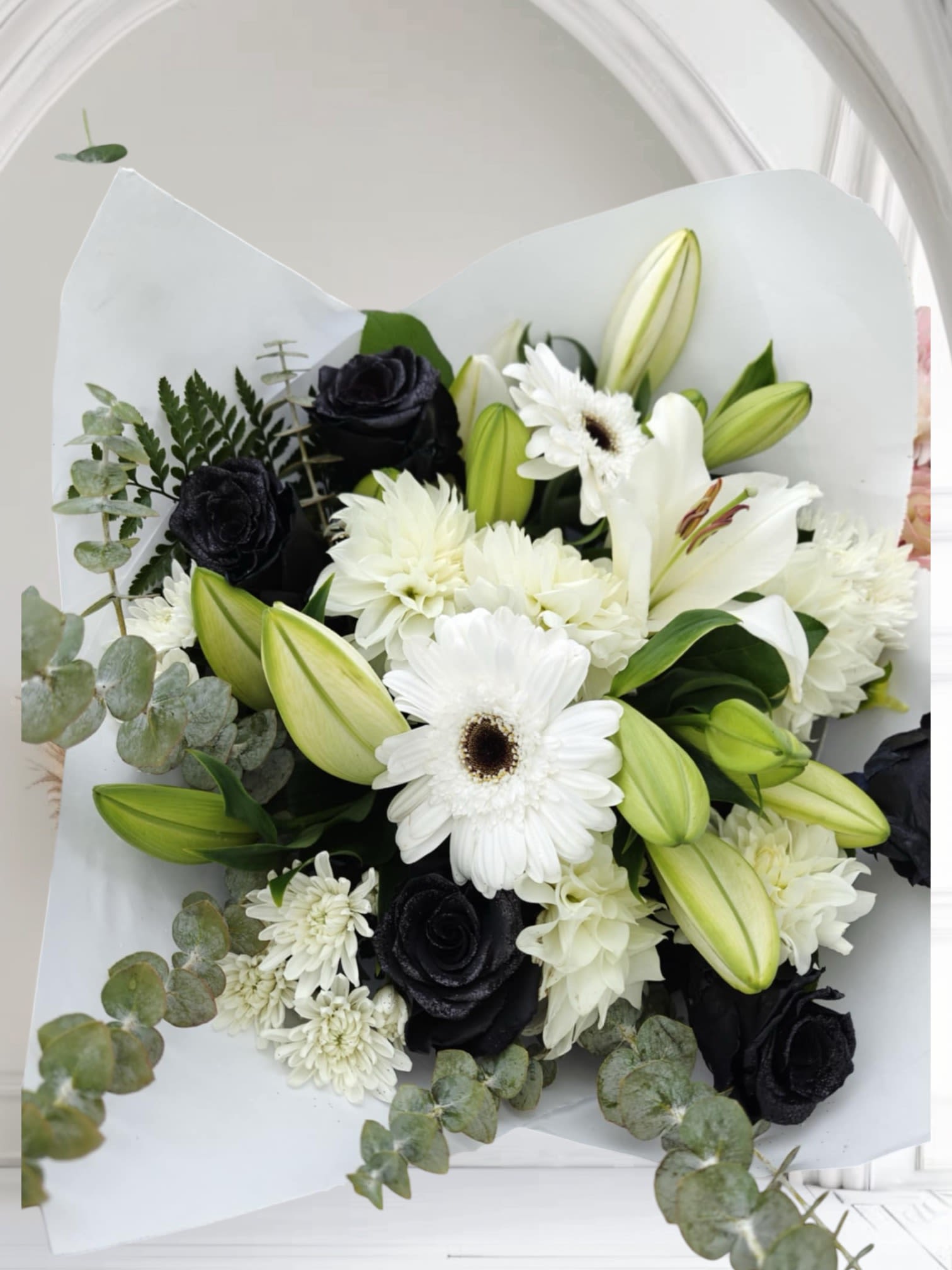Elegant Contrast -  This bouquet combines striking black roses (rose color may vary )with pure white lilies and Gerber daisies, creating a dramatic and refined look. Soft eucalyptus leaves and ferns add subtle texture, enhancing its timeless elegance.