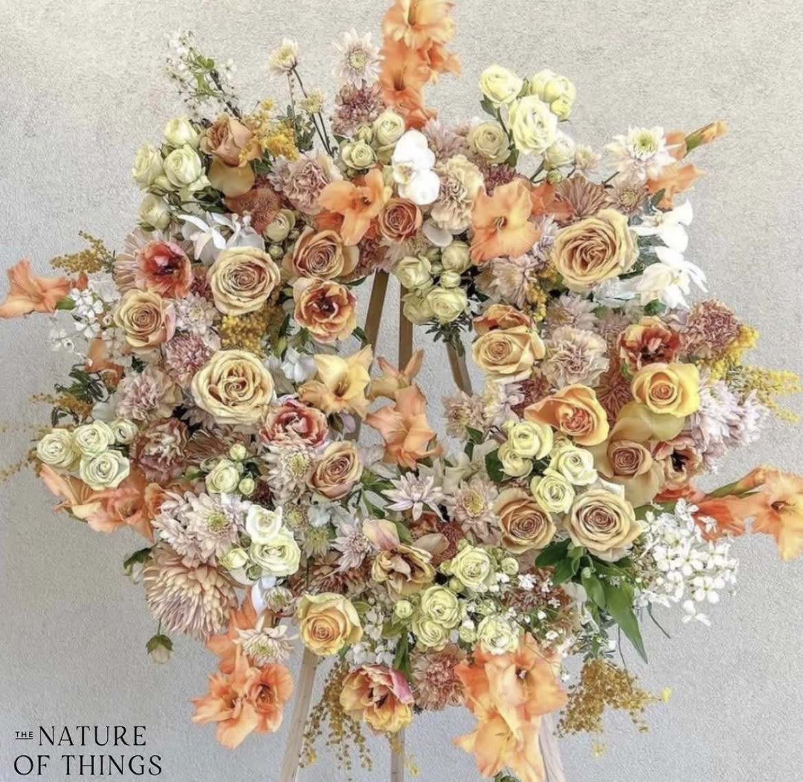 Peach Tones Memorial Wreath - Circular wreath in shades of soft orange, toffee, blush, soft yellow, peach, with touches of white. The season’s most beautiful florals and greenery are featured beautifully in a natural, garden style on a standing easel.  ————————————  Flowers and greenery may vary due to seasonal availability, but that’s part of the charm! Our creative designers will substitute with the freshest high end product available- some grown in our very own garden!- and create something beautiful.  *Have a special request?* Let us know in the special instructions section during checkout.