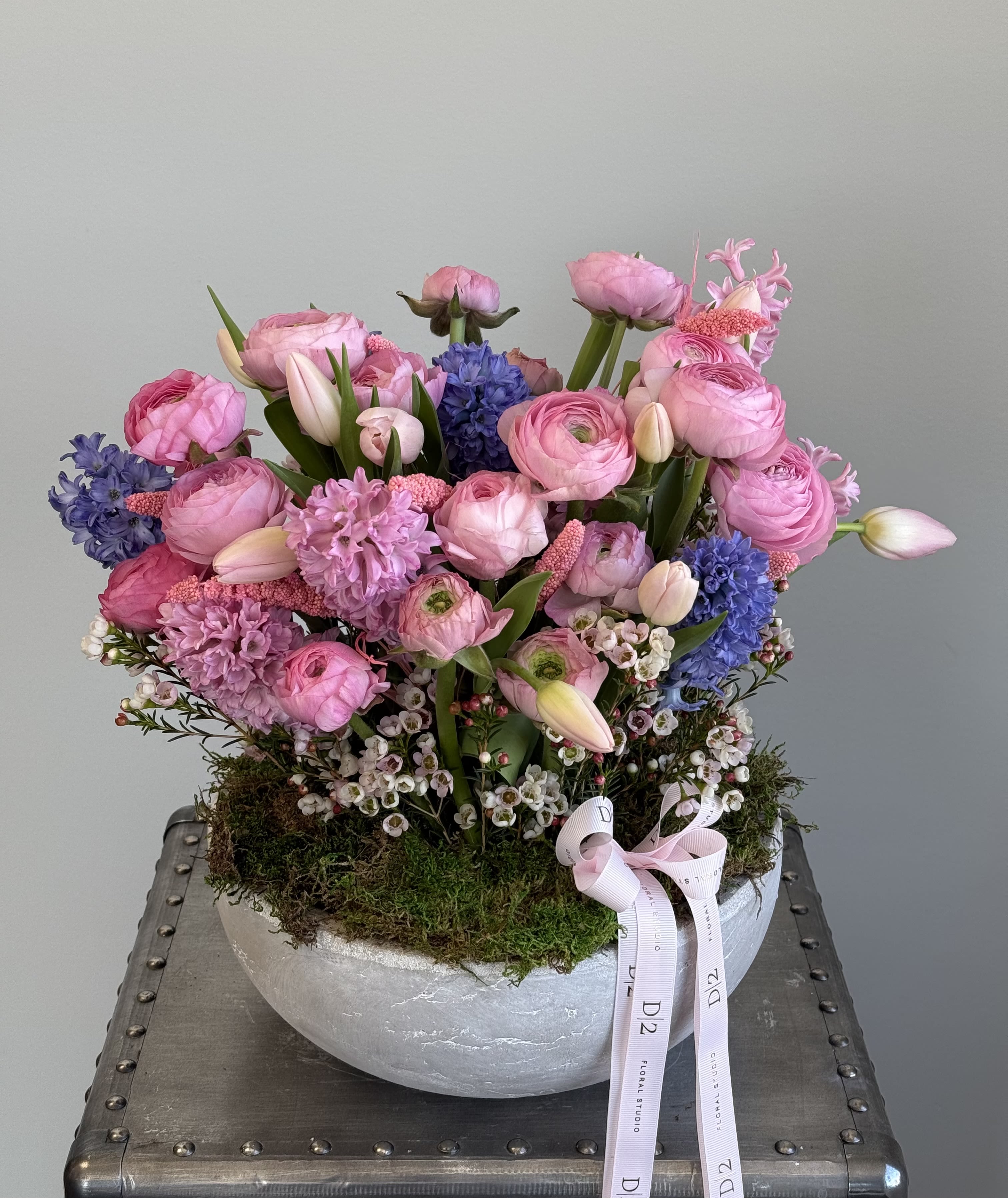 Lidia Lidia - This is a beautifully arranged floral composition featuring a mix of pink and purple blooms. The arrangement includes delicate ranunculus, tulips, hyacinths, and wax flowers, complemented by lush greenery and moss at the base. It is presented in a modern, textured stone-like bowl with a soft ribbon accent, giving it an elegant and sophisticated feel. The combination of pastel tones and fresh, seasonal flowers creates a romantic and timeless aesthetic.