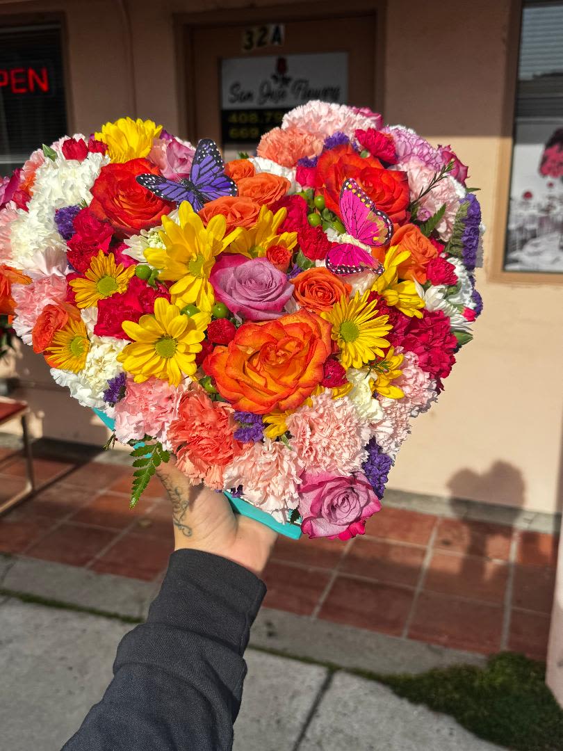 Springtime Love Blooms - A stunning heart-shaped bouquet featuring a colorful mix of flowers, including roses, carnations, daisies, and other blooms in shades of orange, pink, yellow, red, and purple. Delicate butterfly decorations add a whimsical touch to this beautifully arranged floral display. The bouquet is held outdoors in front of a flower shop, enhancing its natural beauty.