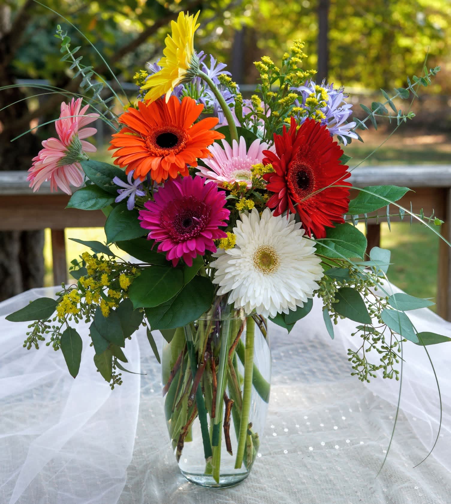 Karen's Kindness - One of our Weekly Wonders and Beautimous originals, Karen's Kindness is perfect for either someone who has shown kindness or if you want to spread a little kindness in the world. These mixed colored Gerbera daisies are surrounded by blue agapanthus, yellow solidago, lily grass, salal, seeded eucalyptus in a tall, clear vase. This carrier of kindness is perfect for all occasions!   APPROXIMATE DIMENSIONS - 24&quot; in  x 12&quot; in     ow