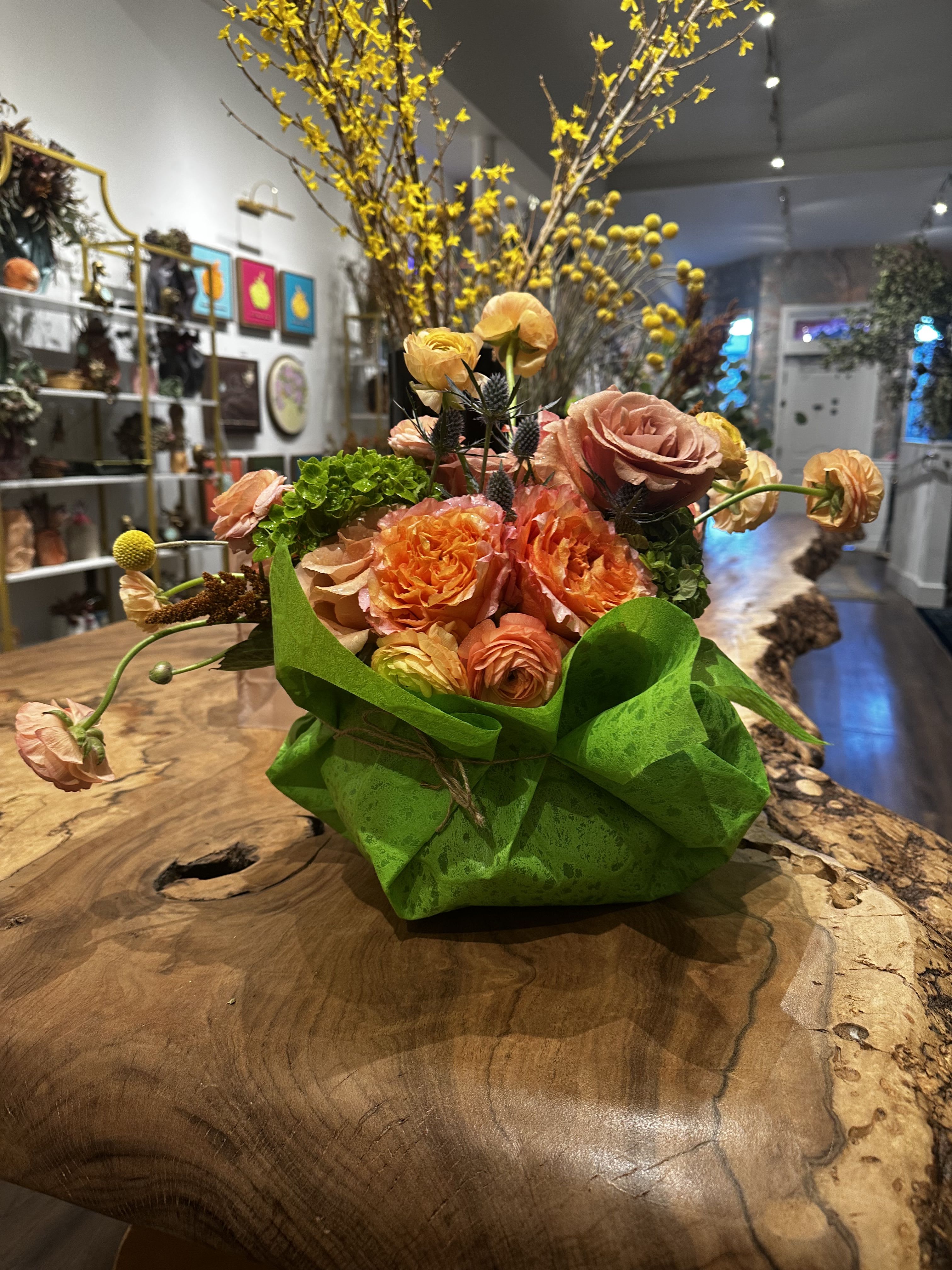 Rococo Glam Arrangement - A whimsical celebration of nature's extravagance, where coral-hued Free Spirit garden roses unfurl their petals in dramatic sweeps reminiscent of 18th-century opulence. Dancing ranunculus in shades of peachy amber create playful movement throughout, while clusters of verdant hydrangea provide a lush foundation. Sculptural blue thistle adds an avant-garde touch, their spiky silhouettes creating beautiful tension against the soft petals. Cheerful billy balls dot the arrangement like golden jewels, adding a modern twist to this baroque-inspired design.  Artfully composed in a sleek glass bowl, the arrangement embodies the Rococo period's love of ornate beauty and theatrical flair. Each stem curves and twirls with artistic abandon, creating an asymmetrical masterpiece that catches light and shadow. The transparent vessel provides a contemporary stage for this romantic performance of florals.  Perfect for those who appreciate the grandeur of classical design with a playful, modern spirit. This arrangement brings a touch of artistic drama to any space while maintaining an air of sophisticated whimsy.  