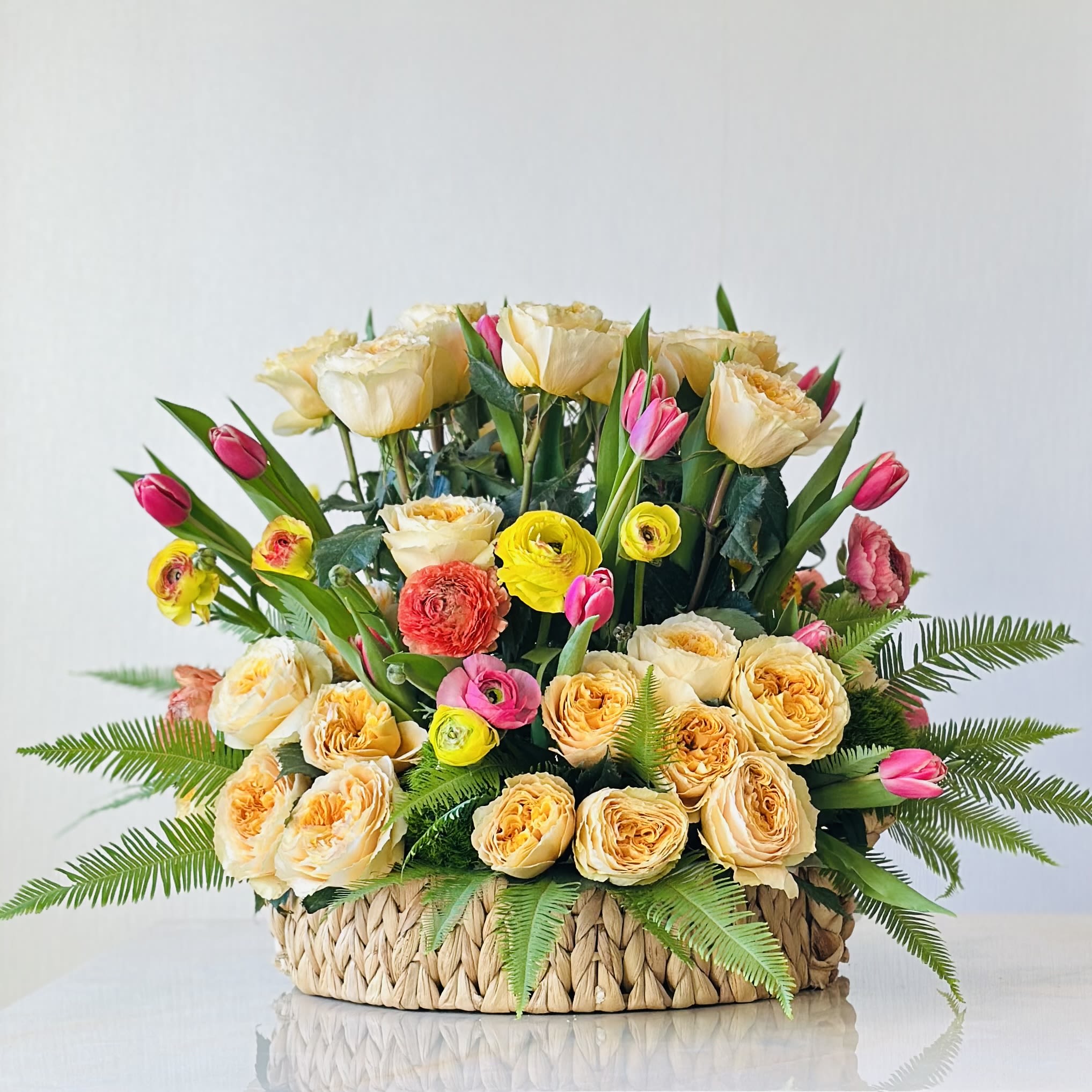 Sunlit Garden Sunlit Garden - Brighten any space with our stunning Sunlit Garden arrangement. This vibrant display features lush yellow roses, delicate pink tulips, and cheerful ranunculus blooms, all nestled in a woven basket for natural, rustic touch. Soft ferns add texture and balance, creating a design that feels both lively and elegant. Perfect for celebrations, special occasions, or simply bringing warmth and joy into your home.