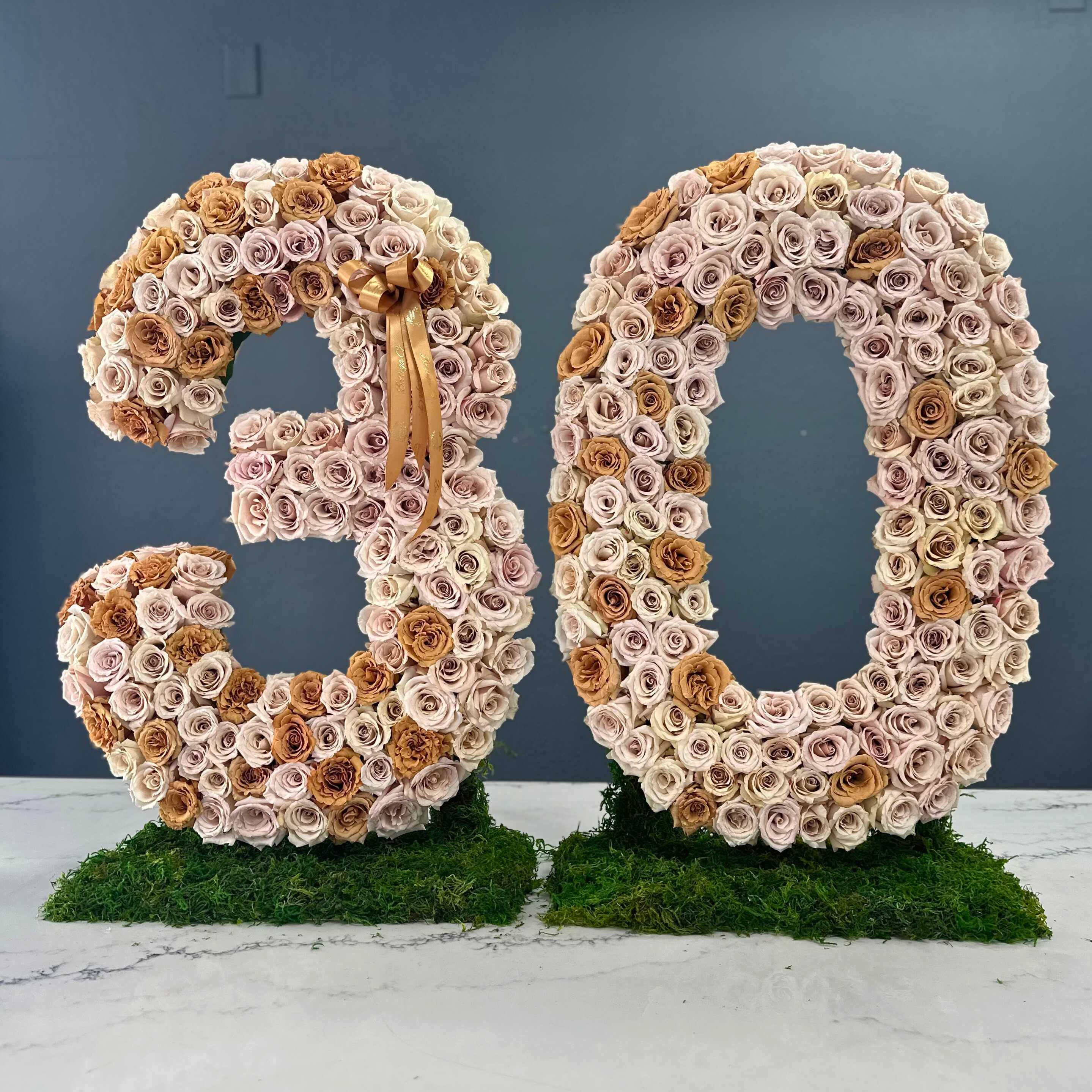 Toffee Tango: A Floral Celebration - Marking three decades of love, laughter, and memories, our Timeless Thirtieth floral arrangement is a tribute to the enduring bond you share. Nestled among lush greenery, the focal point of this arrangement is the elegant Quick Sand roses, their dusky pink petals symbolizing the depth and warmth of your journey together.  Intertwined with the Quick Sand roses are Toffee roses, their rich caramel tones adding a touch of sweetness to the ensemble, reminiscent of the cherished moments you've shared.  Amidst the blooms, a delicate number &quot;30&quot; stands tall, crafted from slender twigs and adorned with tiny blossoms, serving as a visual reminder of the milestone you've reached.  As an additional touch, scattered throughout the arrangement are pearl accents, representing the precious pearls of wisdom gained over thirty years of shared experiences.  In the notes accompanying this arrangement, let us know any specific number you'd like to include, perhaps significant dates or milestones, as we customize this floral masterpiece to reflect your unique journey.       