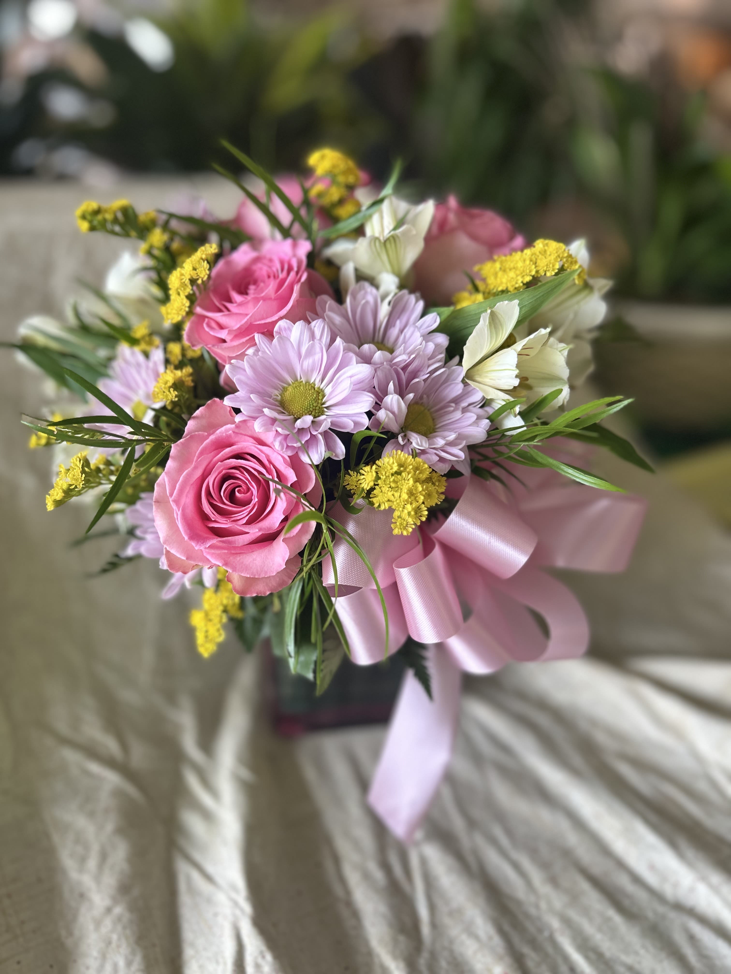 Spring Blossoms -  A beautiful and vibrant spring arrangement! Imagine a soft pink cube filled with a mix of delicate pink roses, white alstroemeria, cheerful purple daisies, and a pop of yellow limonium to add a playful, sun-kissed touch. It  definitely brings a refreshing, breezy feeling of spring, capturing both the elegance and the joy of the season. The contrast of the colors would feel lively and full of warmth!