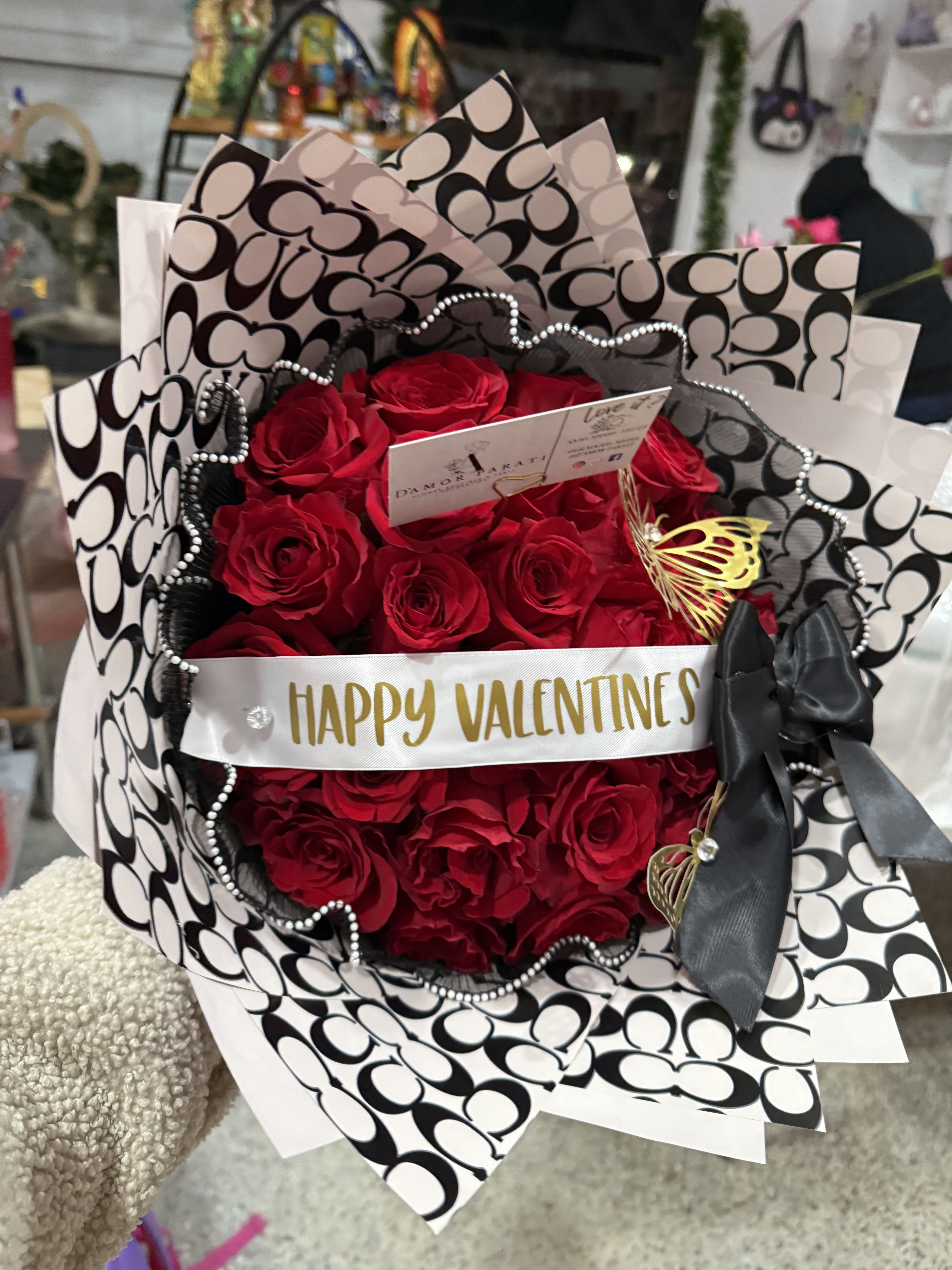 Red Valentines Red Valentines - A 25 count bouquet of red roses is wrapped in Coach-patterned paper and a white ribbon with "Happy Valentines" written on it. The roses are a deep red color, with varying shades and textures suggesting a realistic or high-quality artificial flower arrangement. The Coach-patterned paper is a black and white design, adding a stylish and recognizable pattern to the presentation. The white ribbon, which is centrally located and slightly elevated above the bouquet, features the text "Happy Valentines" in gold lettering. Small, decorative elements like a gold butterfly embellishment and a black bow add further detail. The overall presentation is neat and visually appealing. The gold butterfly is a smaller decorative element that adds a delicate touch to the arrangement. The black bow adds a touch of elegance and formality to the presentation.
