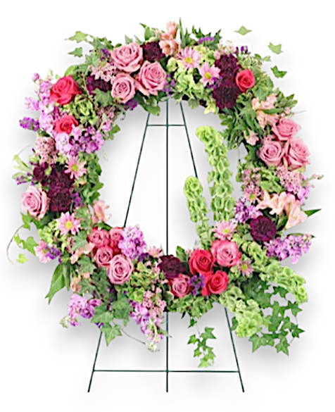 Ever Enchanting Easel Ever Enchanting Easel - Elegantly displayed on a wire easel, this striking sympathy arrangement showcases a large wet floral foam wreath, designed to preserve the freshness of its vibrant blooms. A lush variety of foliage, including variegated pittosporum and delicate ivy, creates a harmonious backdrop that enhances the wreath’s eye-catching design. At its center, 13 exquisite lavender roses and bold hot pink roses harmoniously blend to convey a message of love and remembrance. Accentuating the arrangement are fragrant stems of lavender stock, carefully cut green hydrangea, and a beautiful array of pink alstroemeria and stems of pink waxflower, adding depth and texture. The cheerful presence of bells of Ireland and rich purple carnations, along with stems of lavender daisy poms, lend an air of elegance and gratitude, making this wreath a heartfelt tribute that beautifully honors the memory of a loved one.