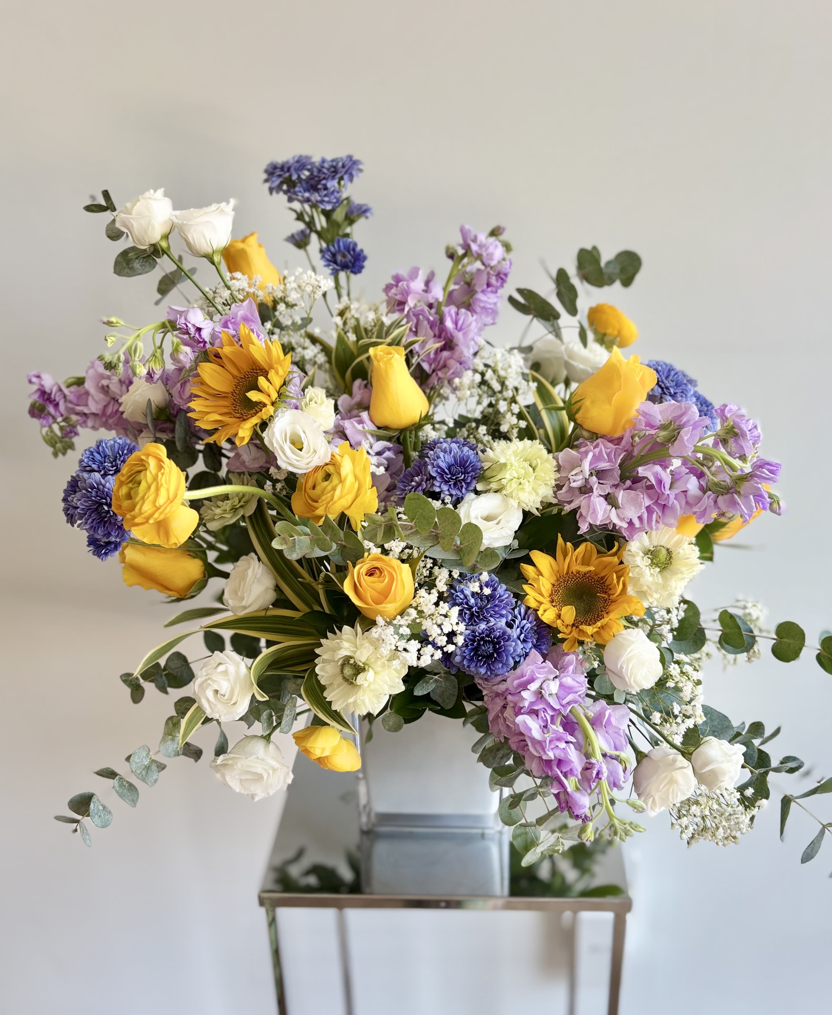 Flora - An asymmetrical, artfully crafted floral piece for special occasions you celebrate or commemorate. In the white-yellow-blue/lavender color palette, the premium flowers are arranged in a 5inch white glass cube. The selection of flowers may vary based on the availability, usually roses, stock, delphinium, sunflowers, ranunculus, lisianthus, anemones, tulips.