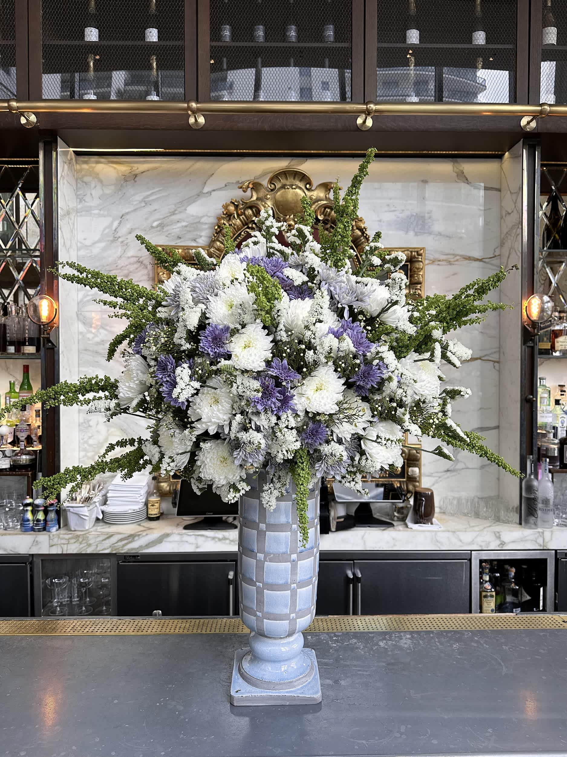 Grand Lavender Elegance Vase - Elevate your space with the &quot;Grand Lavender Elegance Vase.&quot; This magnificent arrangement features an exquisite blend of white and lavender chrysanthemums, accented with delicate green fillers and lush greenery. Arranged in a tall, stately vase, this piece brings an air of sophistication and grandeur to any setting. Please note, vase styles may vary slightly – contact us for current options.  Arrangement Details:  Main Flowers: Chrysanthemums, Green Fillers, Premium Blooms Container: Tall artistic vase (styles may vary, please call for current options) Approximate Dimensions: 24&quot;D x 48&quot;H