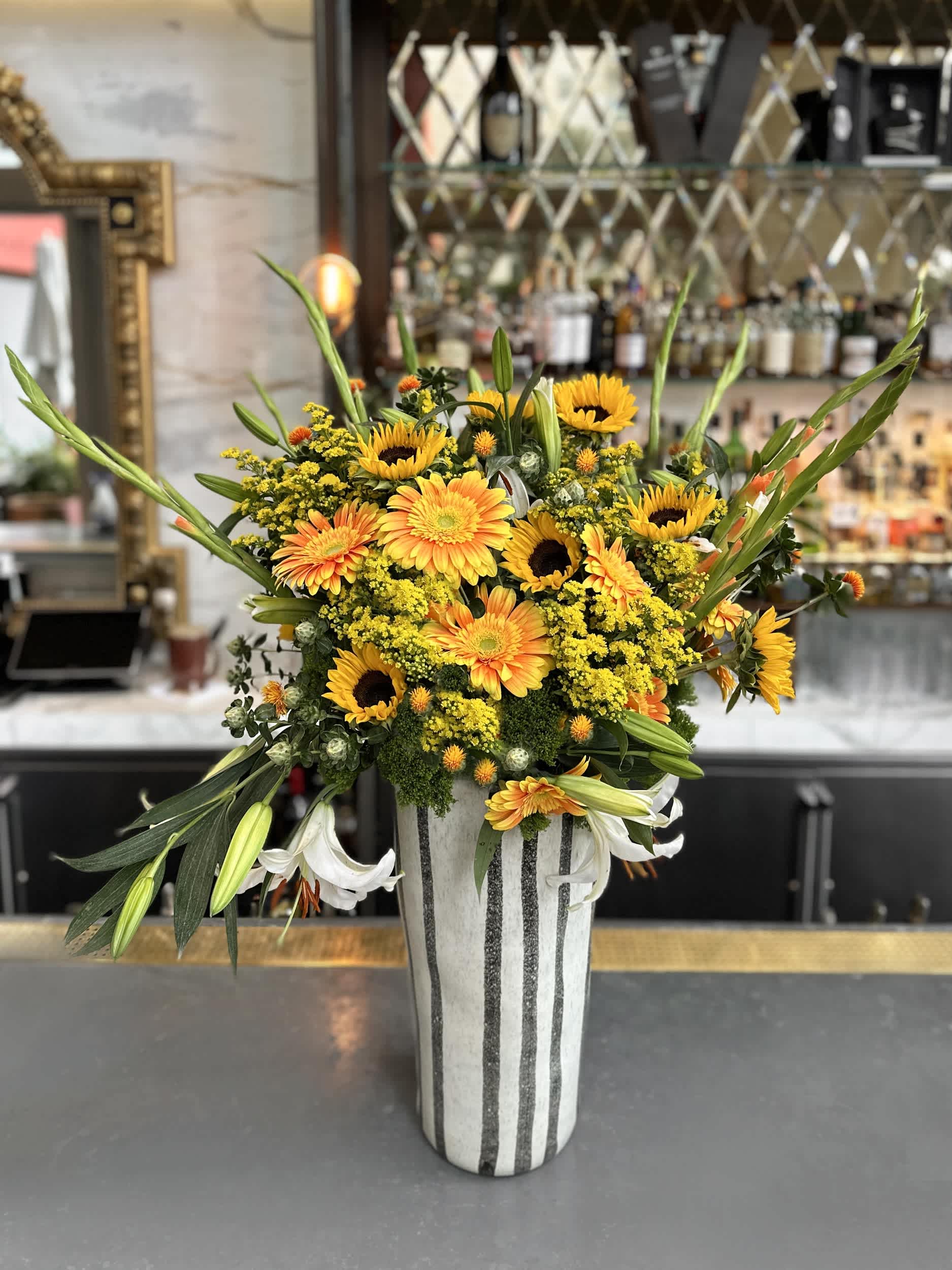 Grand Sunshine Radiance Vase - Bring a burst of sunshine into any room with the &quot;Grand Sunshine Radiance Vase.&quot; This vibrant arrangement features a mix of sunflowers, gerbera daisies, and lilies, complemented by lush greenery and goldenrod for a touch of warmth and brightness. Arranged in an artistic striped vase, this design is perfect for making a bold statement at any event or as a stunning centerpiece.  Arrangement Details: Main Flowers: Sunflowers, Gerbera Daisies, Lilies, Goldenrod, Greenery Container: Artistic striped vase Approximate Dimensions: 18&quot;D x 36&quot;H