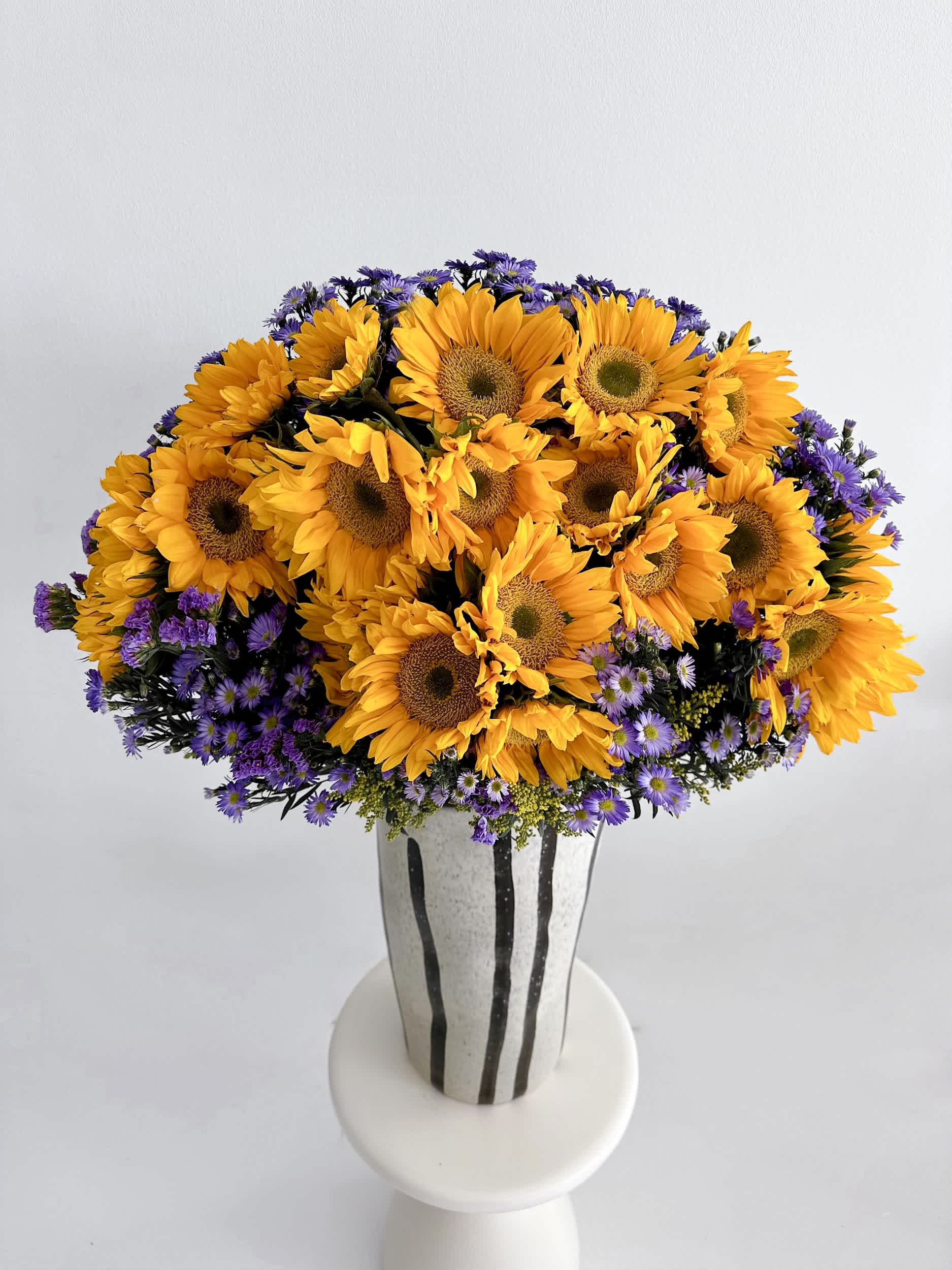 Grand Sunflower Radiance Vase - Brighten any room with the &quot;Grand Sunflower Radiance Vase,&quot; a vibrant arrangement that captures the joyful essence of sunflowers paired with delicate purple asters. This stunning composition brings warmth and cheer, making it the perfect addition to any celebration or to simply add a touch of sunshine to your day. Set in a unique, artistic vase, this arrangement stands out as a beautiful centerpiece.  Arrangement Details: Main Flowers: Sunflowers, Purple Asters Container: Artistic striped vase Approximate Dimensions: 18&quot;D x 36&quot;H