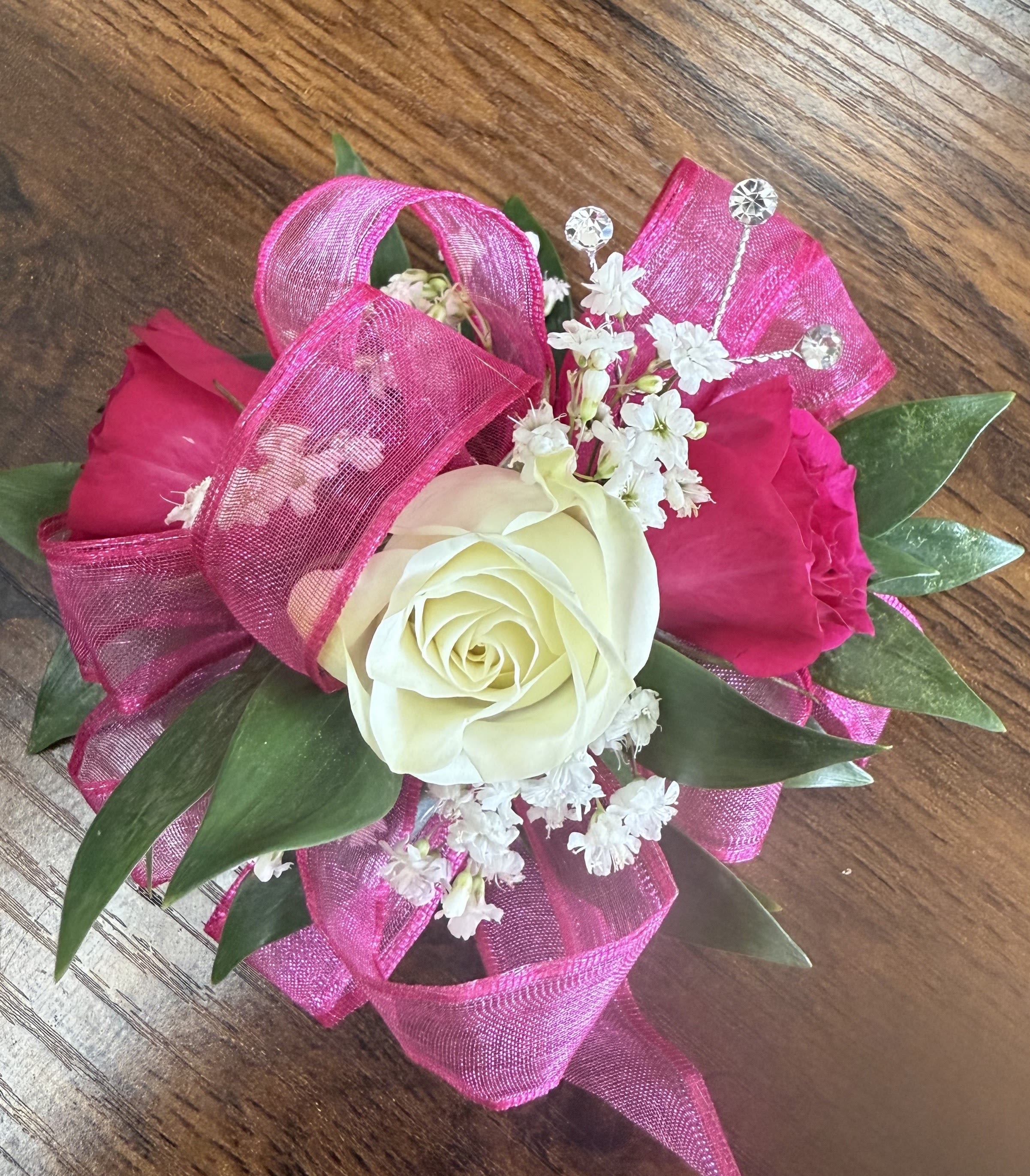 Kid's Hot Pink Corsage - Make her dance unforgettable with our 'Kid's Hot Pink Corsage.' This charming corsage features three delicate spray roses—two vibrant hot pink and one pure white—accented with soft baby's breath, lush greenery, and a sprinkle of sparkling rhinestones. Finished with a flowing hot pink ribbon, it's the perfect touch of elegance for her special night.  For a matching boutonniere, click on the deluxe option!  Measures approximately 4 inches wide and 3 inches high.
