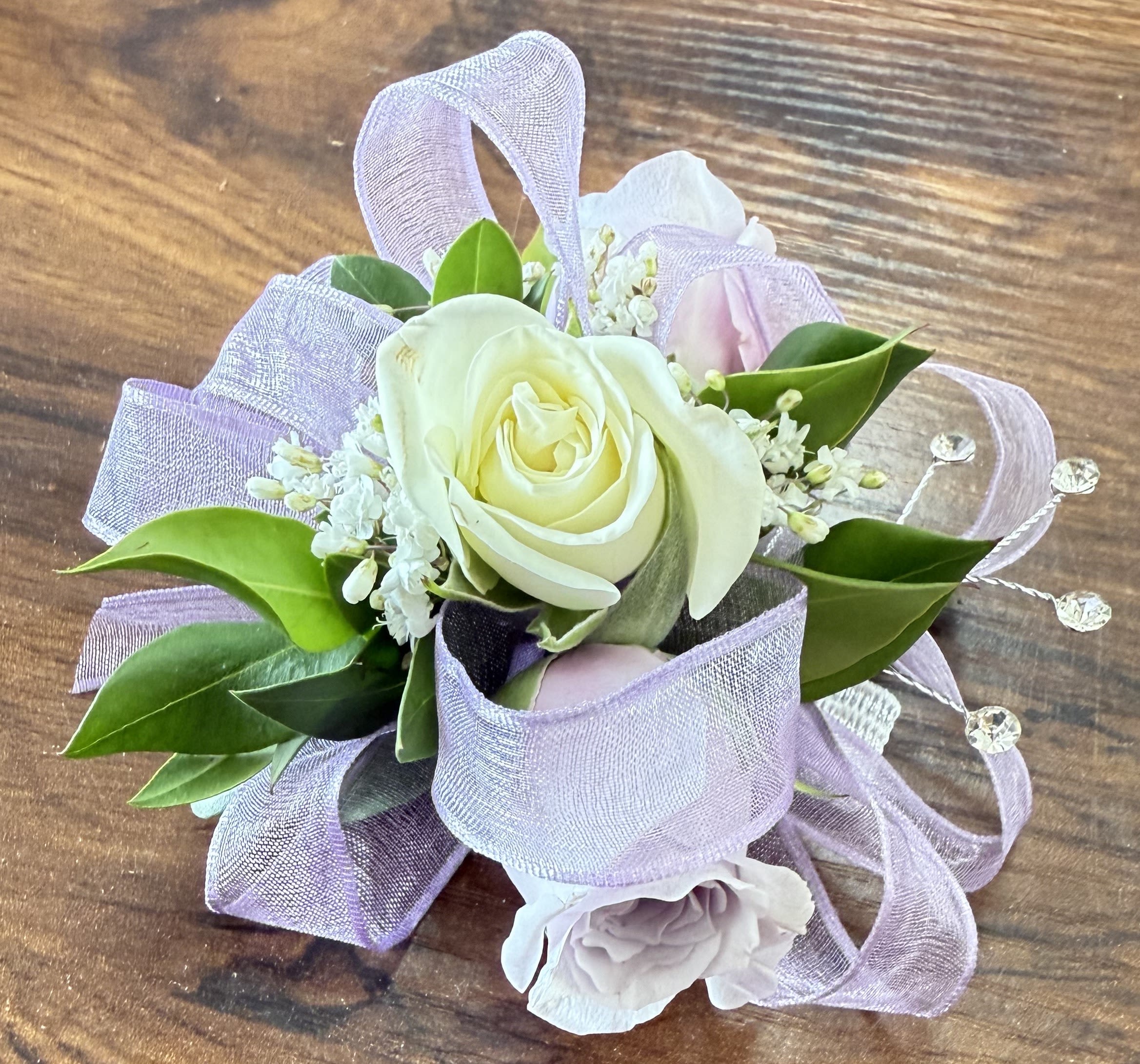 Kid's Lavender Corsage - Let dreams blossom with our 'Kid's Lavender Corsage.' This beautiful wristlet features two lavender and one white spray rose, with baby's breath, greenery, and sparkle. A lavender ribbon completes the magical look. For a matching boutonniere, click on the deluxe option!  Measures approximately 4 inches wide and 3 inches high. 