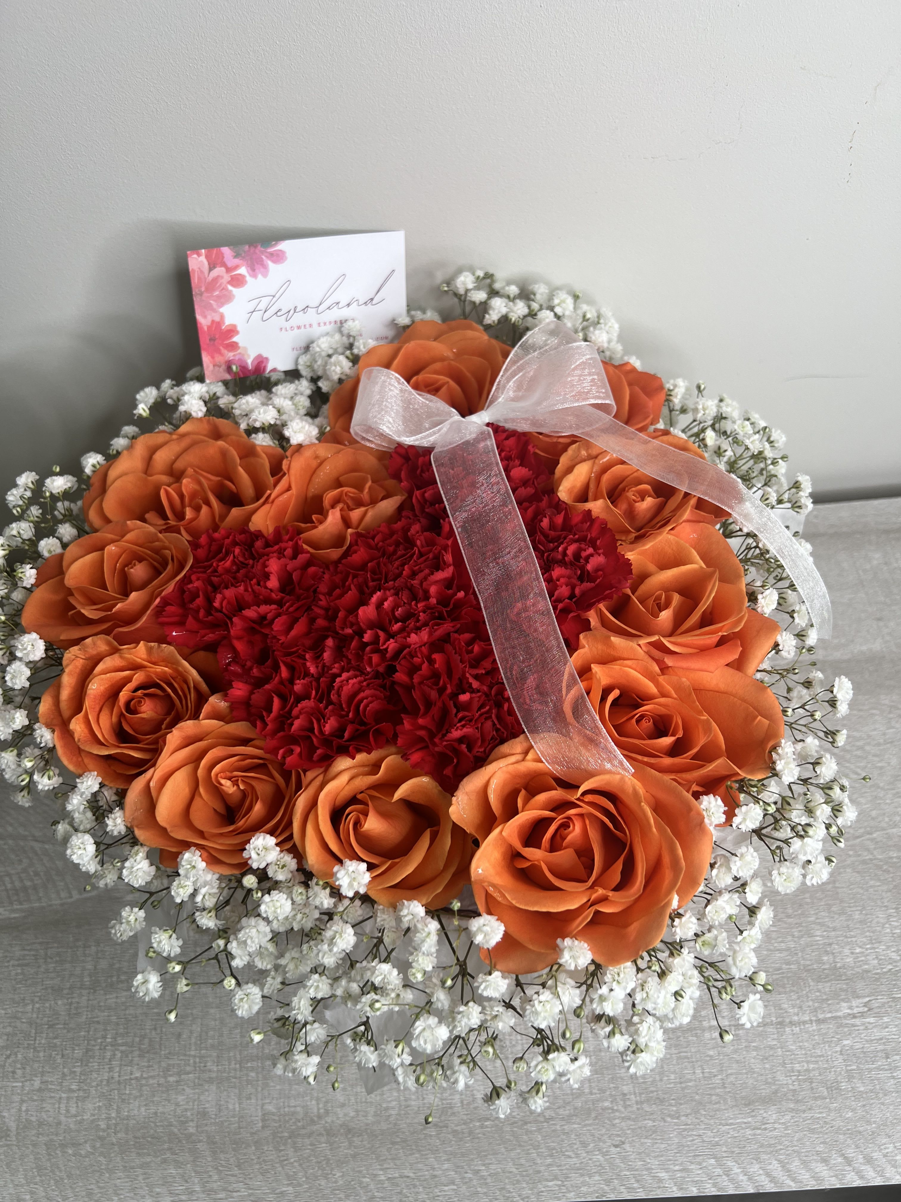 Heart on Fire - This heart-shaped arrangement combines the warmth of orange roses, the intensity of red carnations, and the delicacy of baby’s breath, creating a unique expression of love and admiration. Perfect for a special occasion or to send a heartfelt message.  A vibrant gesture that speaks straight from the heart.