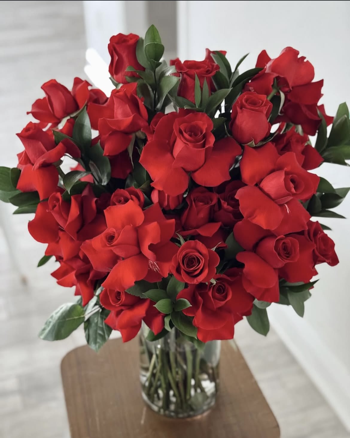Red Burst Rose - This elegant floral arrangement features a lavish bouquet of vibrant red roses, artistically arranged in a clear glass vase. The rich red petals exude romance and passion, while the lush green foliage adds a natural contrast, enhancing the bouquet’s beauty. With its full, symmetrical design and fresh, long-stemmed roses, this classic arrangement is perfect for expressing love, admiration, or celebration in the most timeless way.