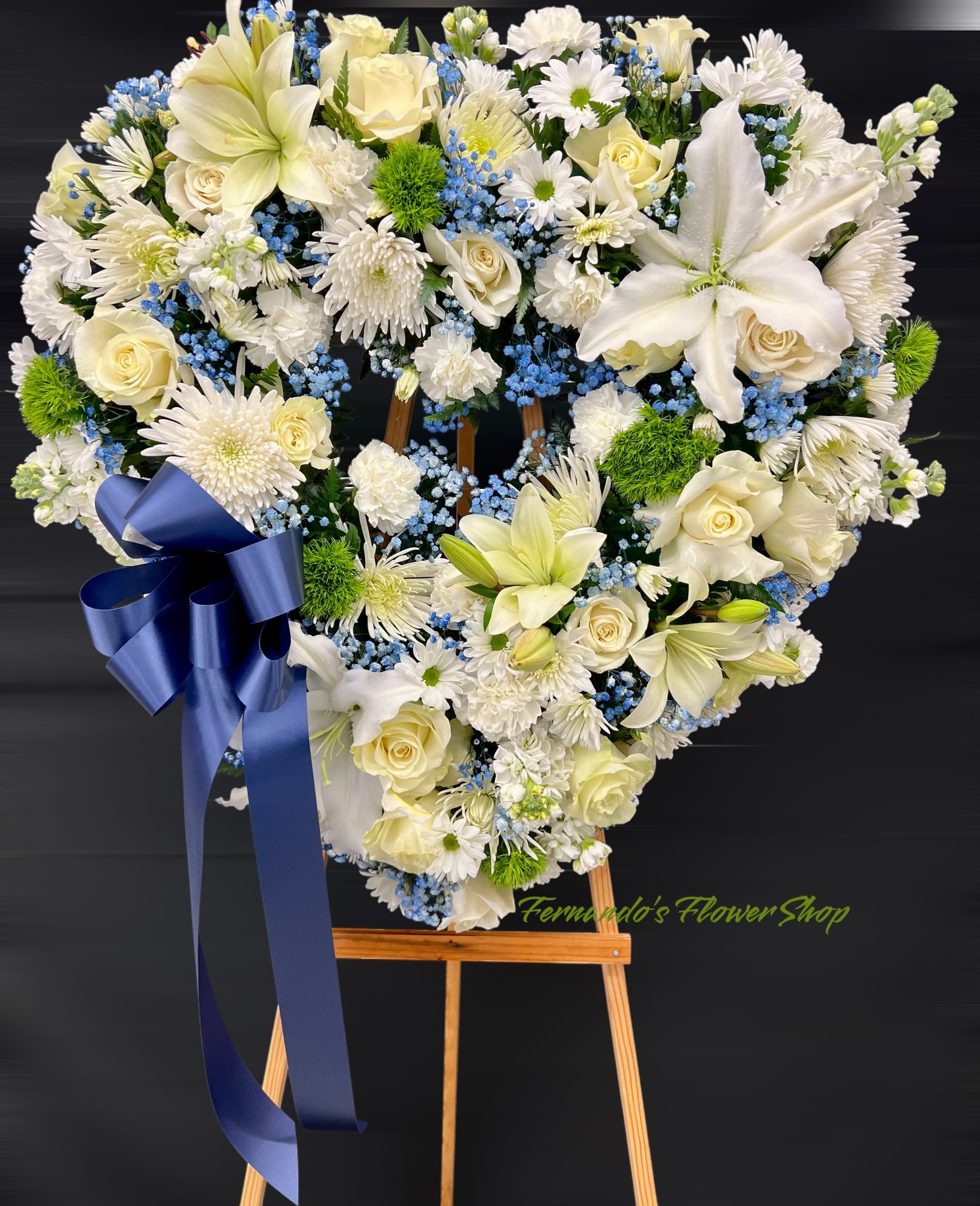 Forever Vanity - This is a heart-shaped floral wreath designed for a funeral or memorial service. It features a mix of white roses, lilies, chrysanthemums, and daisies, complemented by blue baby’s breath and green foliage. A large navy blue ribbon adds a decorative touch, and the arrangement is displayed on a wooden easel. The text “Fernando’s Flower Shop” is subtly placed at the bottom.