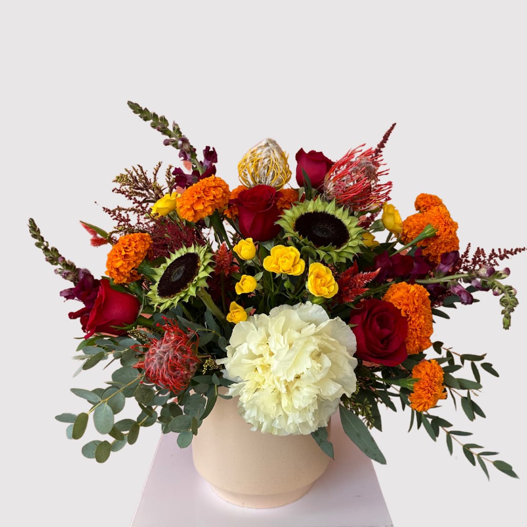 The Lila - The Lila is a radiant celebration of bold color and joyful blooms. Featuring vibrant striped sunflowers, rich red roses, sunny marigolds, striking protea, and soft hydrangea, this arrangement creates a stunning contrast of textures and hues. Perfect for adding a pop of brightness and energy to any occasion, The Lila is designed to bring warmth, cheer, and natural beauty into any space, making it a showstopper wherever it goes.  Please note: The floral design of your piece may be slightly different than the appearance in the images below dependent on the seasonal availability of certain flowers. Thank you for your understanding!
