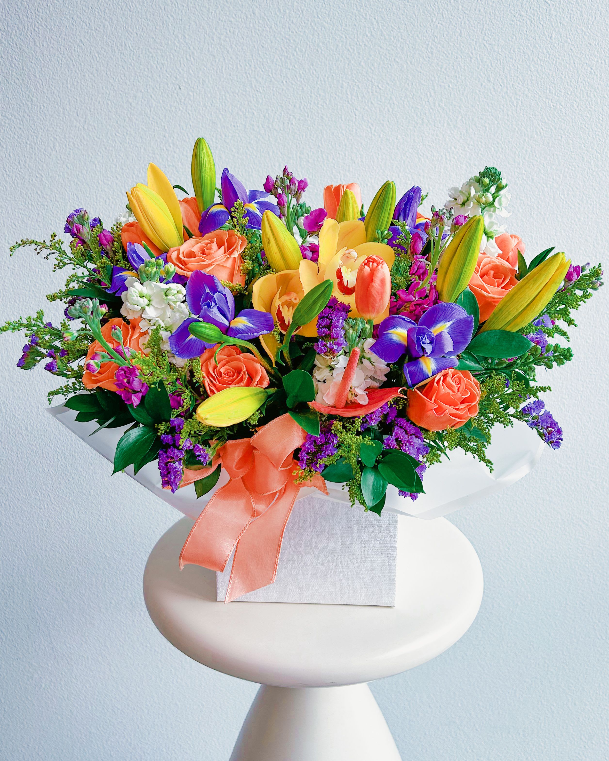 Fancy Flair   - This bouquet is pure sunshine wrapped in a box! With bold yellow lilies, fiery orange roses, and striking purple irises, it’s got that wow factor that makes any space feel alive. The fresh greenery and delicate white stock flowers soften the look, while the bright coral ribbon ties it all together in the most stylish way. Whether you’re celebrating a birthday, saying congrats, or just spreading some happiness, this vibrant beauty is guaranteed to deliver good vibes only!