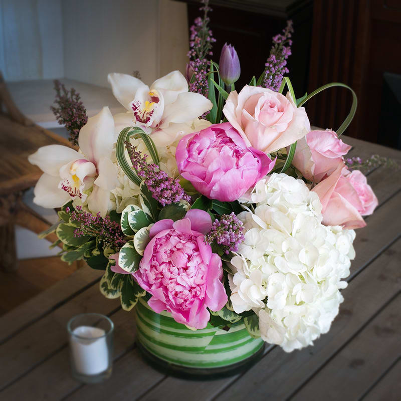 Blush 'N Bloom - A clear glass cylinder vase lined with leaves, concealing the stems of the beautiful flowers arranged inside. This arrangement features Hydrangea, Peonies, Orchids, Roses and Heather decorated with Variegated Lilly Grass and Greens. 