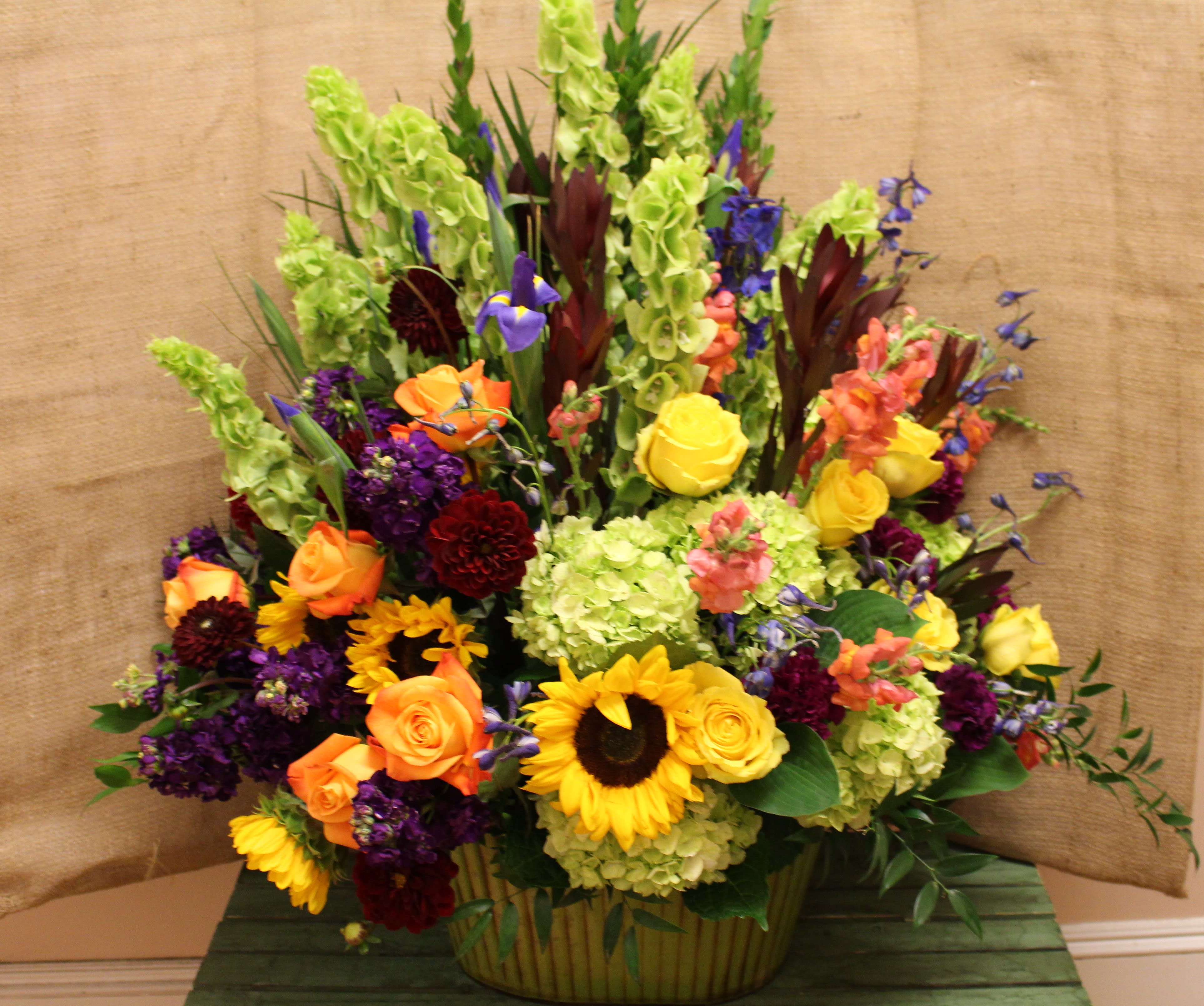 Brazen Beauty  - This green tub is filled with all of our favorite bright flowers to celebrate a loved one. With an impressive display featuring hydrangea, sunflowers, roses, bells of Ireland, stock, carnations, iris, and more, this piece is sure to stand out. With it's bright and vast color line, it makes a sad time just a little easier to bear.