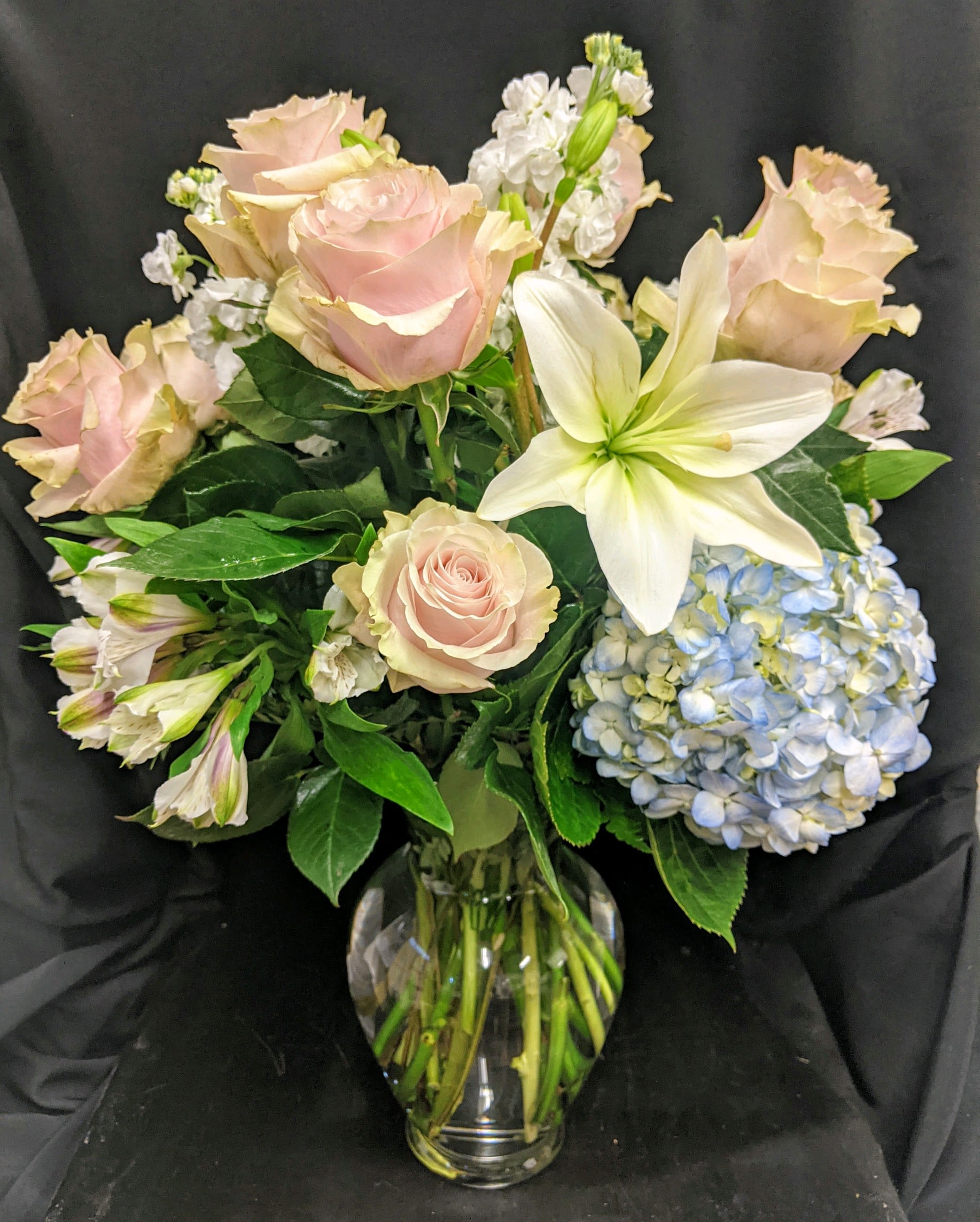 Country Dreamin' (shown in Standard) Country Dreamin' (shown in Standard) - Breathtaking! Blooms of bright white and light pink accented with blue hydrangea and fragrant white stock are rich with texture and innocent beauty. Presented in a clear glass vase. This is a fabulously fragrant choice for a bride-to-be, your beloved mom, or an old friend. • Astonish her by upgrading to deluxe with 9 roses or a full dozen with a premium upgrade. • Light pink roses, white asiatic lilies, white stock, blue hydrangea and beautiful deep green leather are delivered in a ginger jar style vase. • Orientation: All-Around