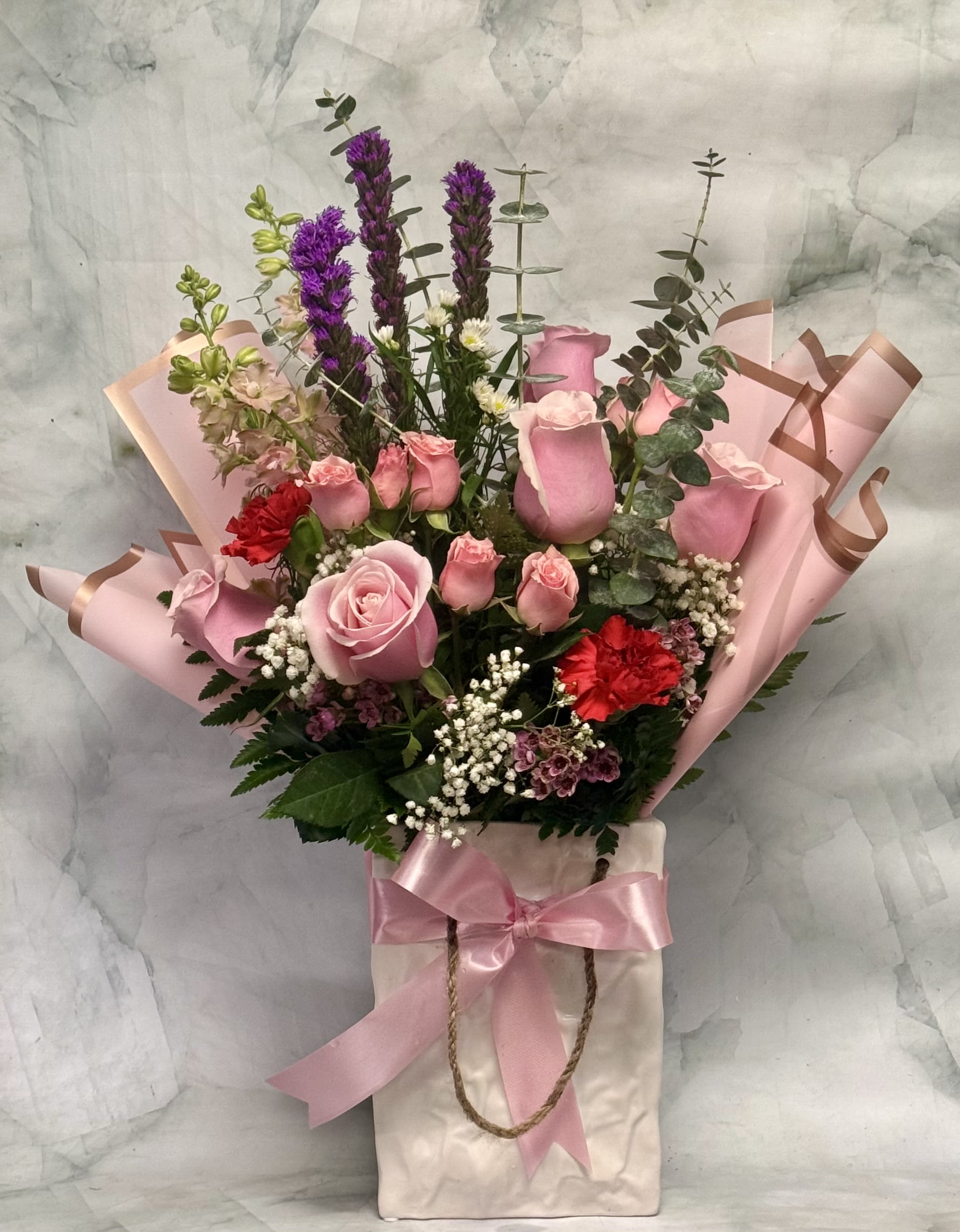 Bag it Up! - This arrangement is already bagged and ready to go. This gorgeous arrangement is designed in our very own white ceramic gift bag container. This adorable keepsake container will give them something to talk about. The arrangement includes roses, spray roses, liatris, larkspur, eucalyptus, wax, babies breath and carnations. 