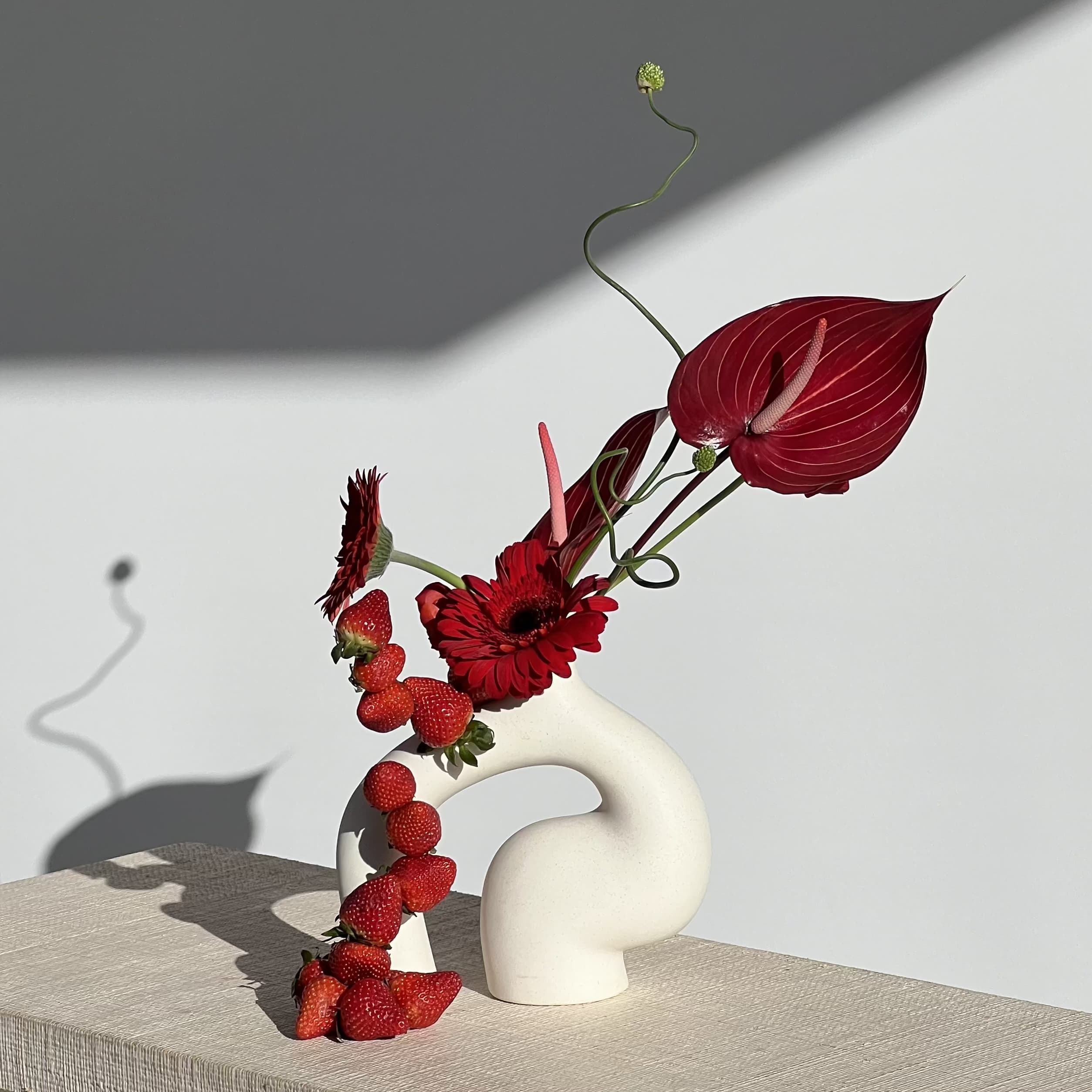 Berry Cute - A minimalist ceramic vase with vibrant red gerbera daisies, accented by a playful cascade of fresh strawberries balancing in front