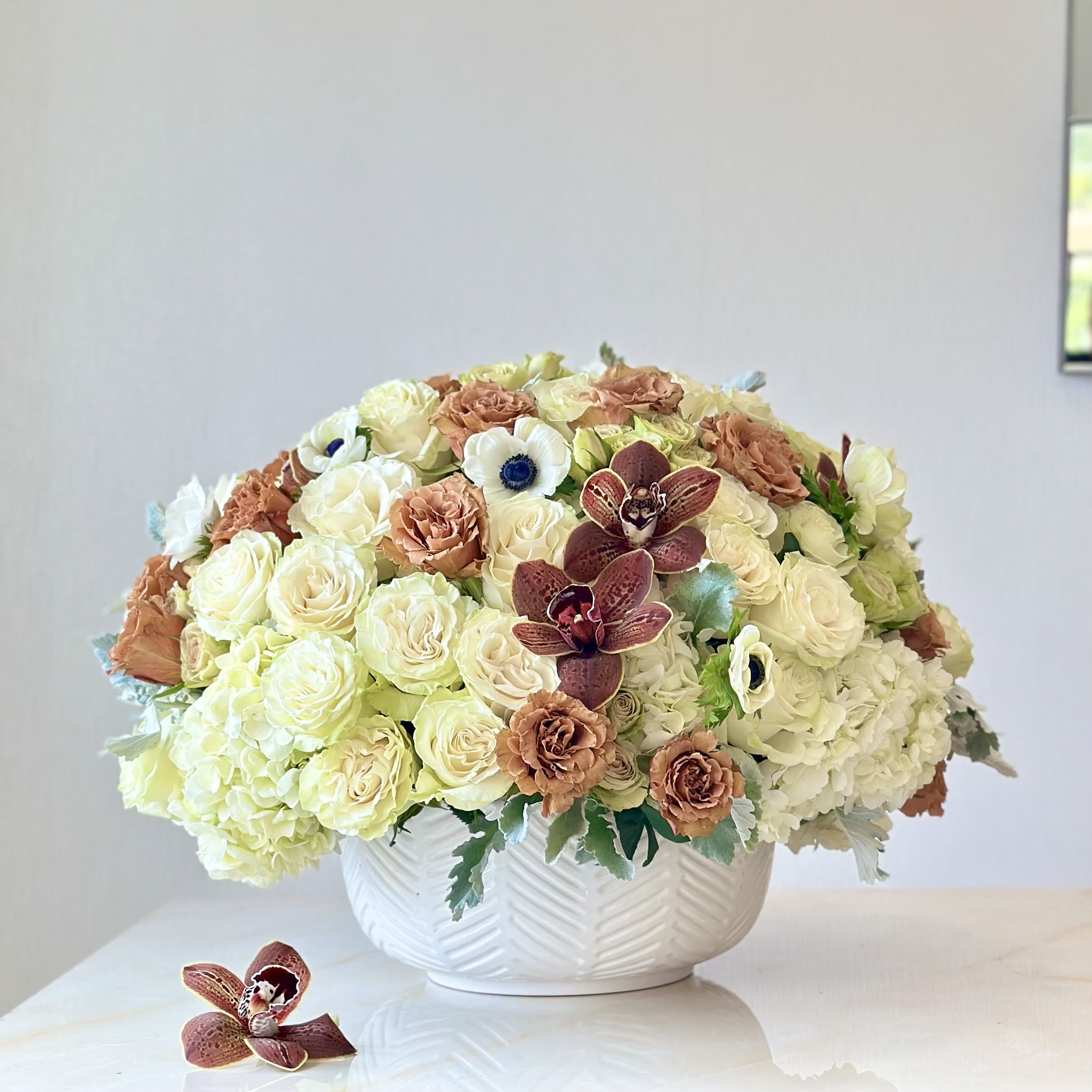 Vintage Glow Vintage Glow - Vintage Glow is a perfect balance of softness and sophistication, blending creamy neutrals with rich caramel tones. The delicate ivory roses and buttery orchids create a timeless elegance, while black-centered anemones add a striking contrast, their dark eyes offering depth and intrigue amidst the softness. Hints of caramel and toffee hues weave through the design, evoking the glow of a late afternoon sun, while wispy foliage adds movement, creating an organic, free-flowing feel. This arrangement is more than an arrangement-it’s a story told texture, tone, and balance.
