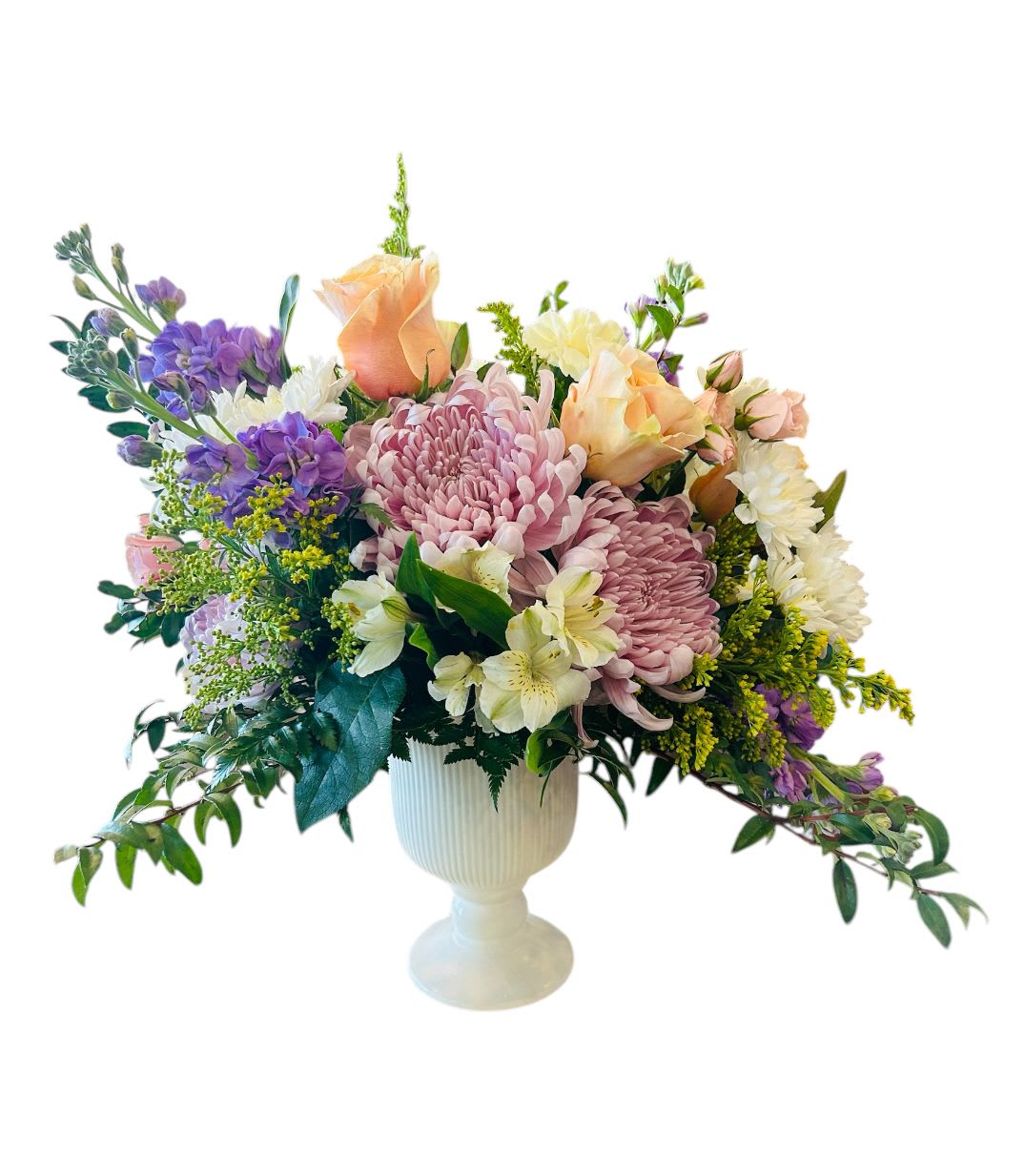 Botanic Bliss - A lush and elegant floral arrangement that combines soft pastels with rich textures, giving it a timeless and romantic appeal. Designed in a classic white pedestal vase, this bouquet features:  *Blush and peach roses – Delicate and soft, adding warmth and romance. *Lavender and cream chrysanthemums – Large, full blooms that bring texture and an antique charm. *White alstroemeria (Peruvian lilies) – Adding a touch of elegance and depth. *Purple stock flowers – Bringing fragrance and a subtle pop of color. *Golden solidago – A touch of yellow to brighten and balance the soft pastel palette. *Greenery with cascading accents – Providing movement and a natural, garden-fresh look.  This arrangement is perfect for everyday, celebrations, or as a statement piece for a classic, refined setting.