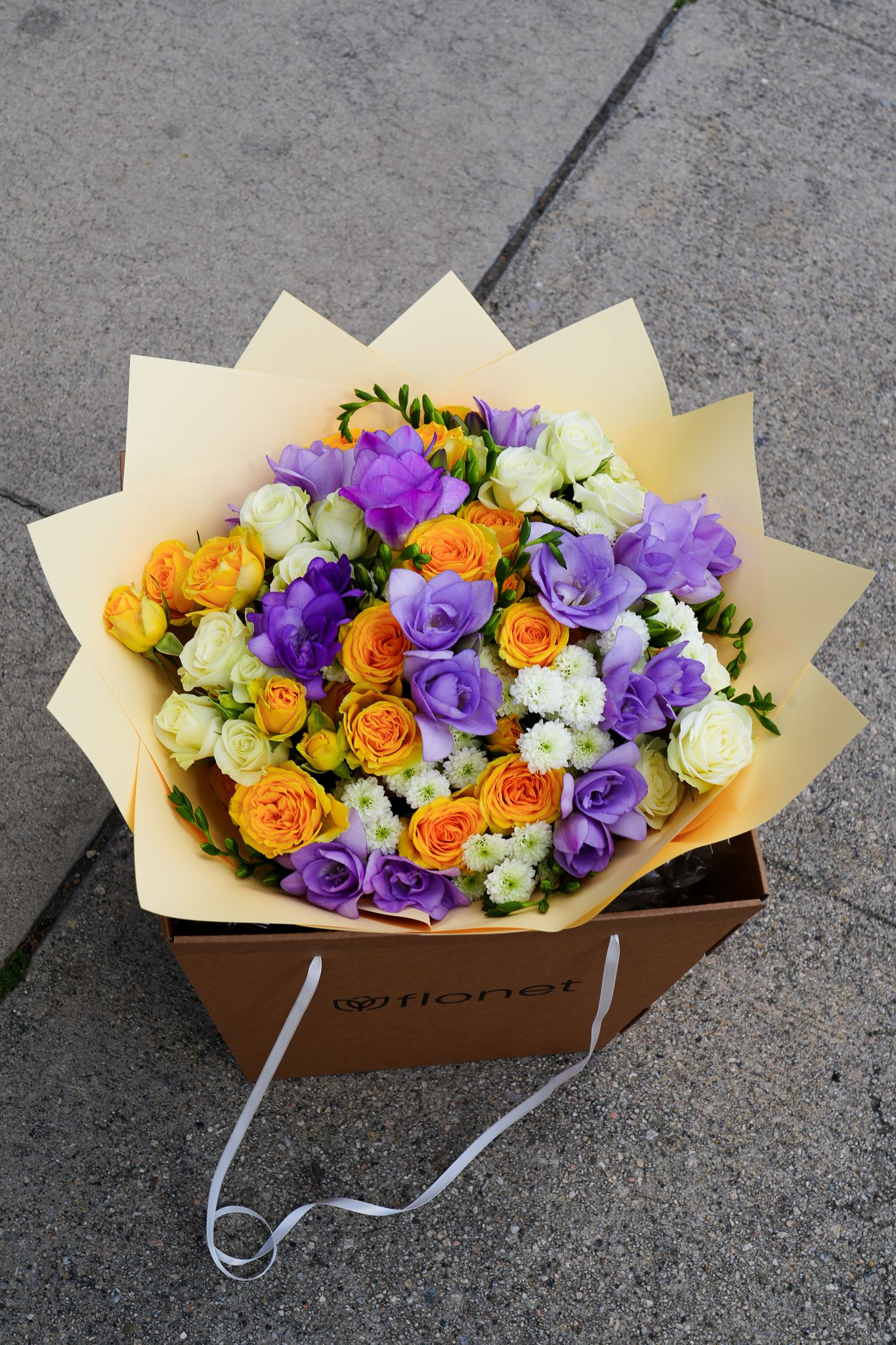 Sunset Symphony - Packaging: Flonet premium kraft bag with satin ribbon handles An elegant celebration of vibrant color and soft charm, Sunset Symphony features a joyful mix of golden yellow and orange roses, lavender freesia, creamy white spray roses, and delicate white chrysanthemums. Wrapped in soft ivory paper and presented in our signature Flonet bag, this bouquet brings warmth, luxury, and a splash of personality to any moment — perfect for birthdays, congratulations, or simply brightening someone’s day.