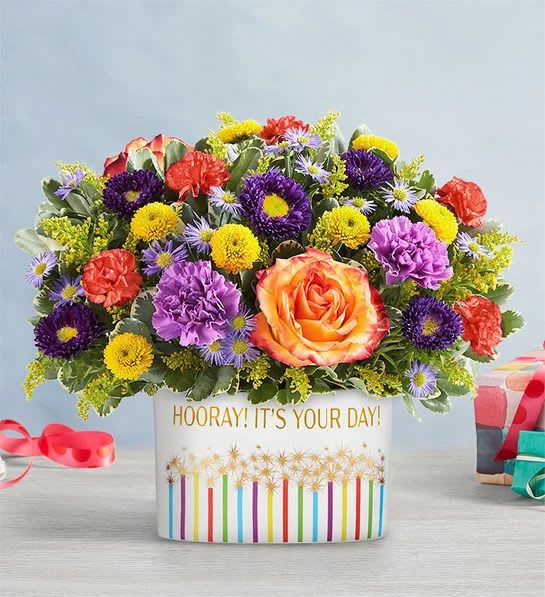 Horray it's your day Horray it's your day - Hooray! It’s Your Day! Floral Arrangement Radiant Roses – A stunning focal point with a vibrant orange and yellow rose, symbolizing joy and admiration. Charming Carnations – Beautiful red and purple carnations add texture and a touch of elegance. Delightful Daisies – Cheerful yellow daisies bring a sunny and uplifting vibe. Lovely Alstroemeria – Soft and delicate, representing friendship and devotion. Monte Casino & Fillers – Accents of purple monte casino and lush greenery enhance the fullness and beauty of the bouquet. Presented in a festive "Hooray! It's Your Day!" container, this arrangement is perfect for birthdays, celebrations, and special moments!