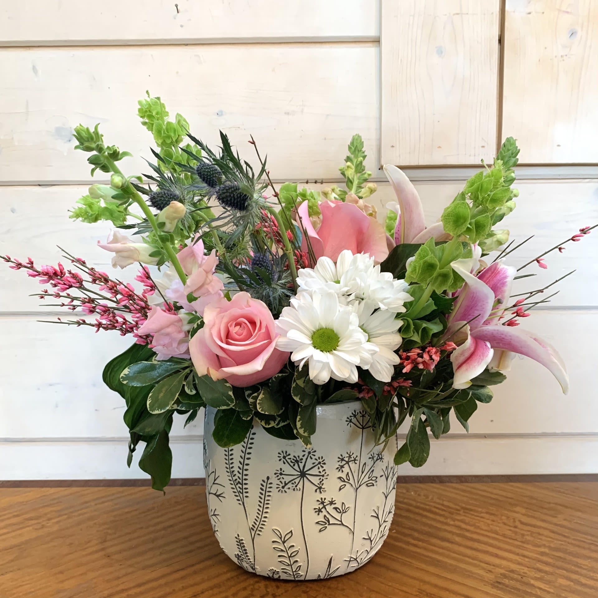 Blushing Blooms Blushing Blooms - Pale pink roses, gazer lilies, pink snaps, green bells of ireland, thistle and seasonal filler flowers arranged in a white ceramic pot embossed with black wildflower designs. Approximately 14in tall and 14 in wide.