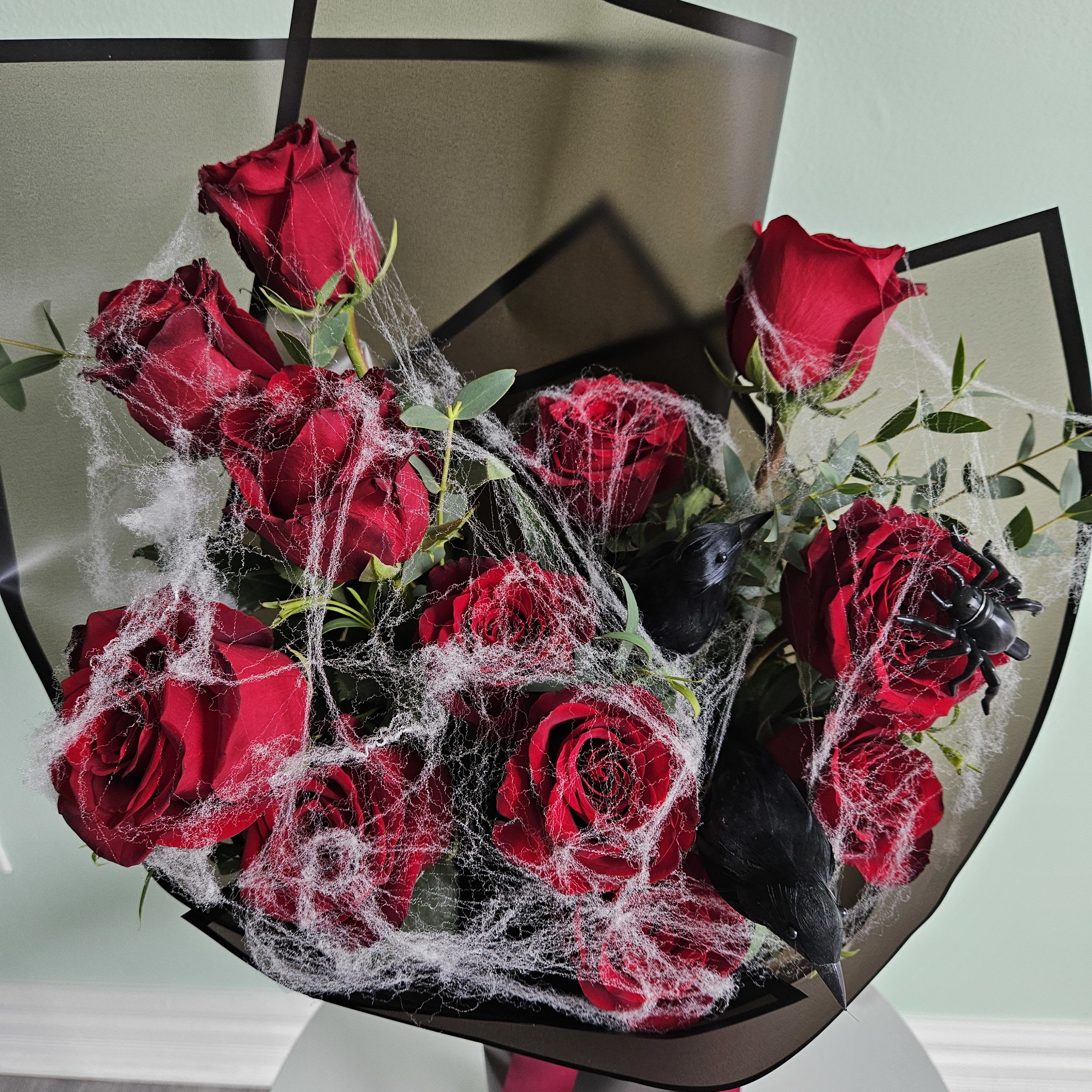 Ravens' Rose Ritual - Our Ravens' Rose Ritual bouquet is a hauntingly beautiful arrangement, perfect for Halloween. Featuring rich red roses wrapped in black paper and covered in eerie spiderweb, it’s complete with two lifelike crows and a spider for a dark, captivating touch. Ideal for adding mystery to your decor or gifting to those who love the spooky season.