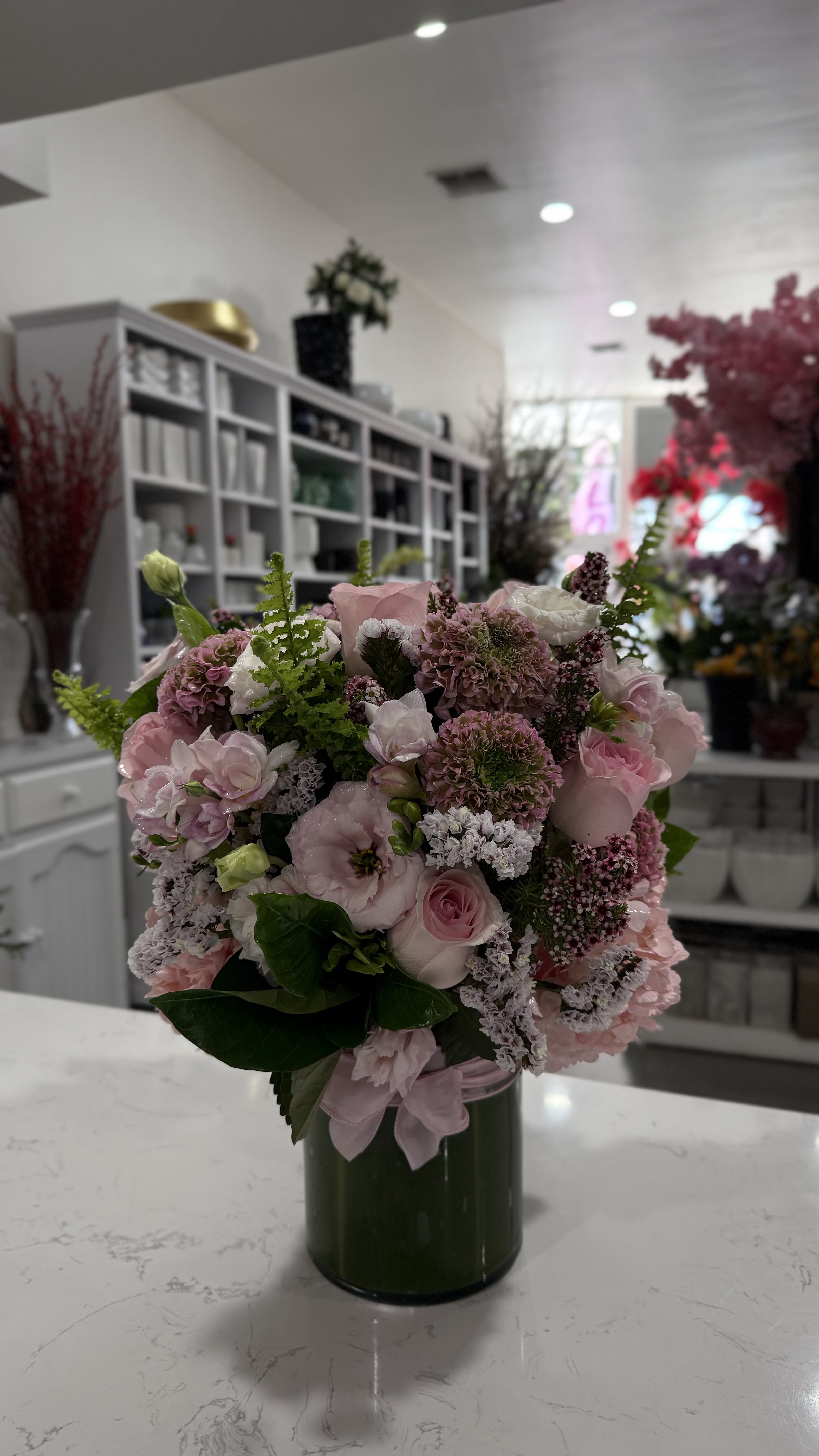 Blush Reverie - A sophisticated blend of soft pink roses, lisianthus, and seasonal blooms accented with textured greens and lavender sprigs. Arranged in a sleek glass cylinder vase, Blush Reverie captures timeless elegance and romantic charm—perfect for celebrations, heartfelt gifts, or simply brightening a space with grace.