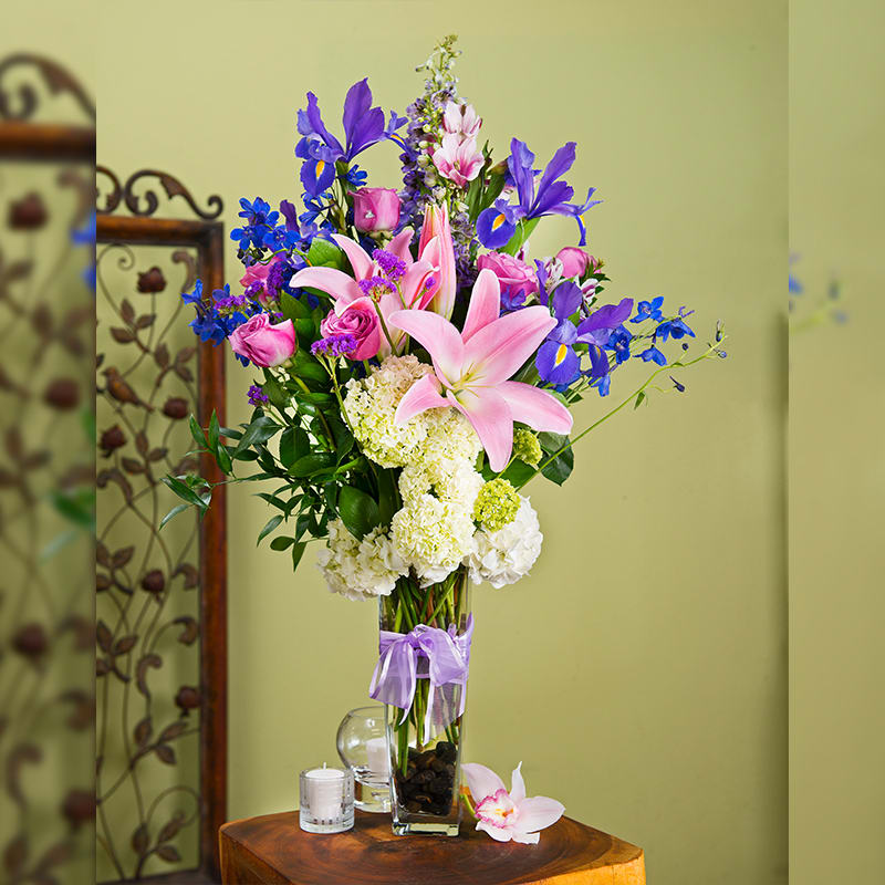 Bobbie - Tall Vase, Purple Flowers, Lillies Iris, Hydrangeas, Lavender roses Bobbie - Tall Vase, Purple Flowers, Lillies Iris, Hydrangeas, Lavender roses - Our tall Special! Spring lavenders are in full bloom and we couldn't be more excited. A tall glass vase with river rock accents, irises, lavender roses, blue delphinium, stargazer lilies, and other assorted purple flowers on a bed of hydrangeas