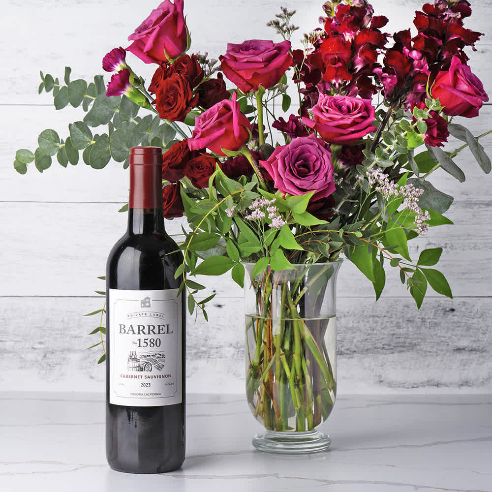 Majestic Rose Arrangement & Cabernet Majestic Rose Arrangement & Cabernet - A luxurious fusion of exquisite blooms and fine wine, perfect for making any occasion unforgettable. This elegant arrangement features a half dozen vibrant magenta roses paired with magenta carnations, creating a rich and captivating color palette. Accented by striking red linear flowers, delicate red spray roses, lush greenery, and charming filler flowers, this bouquet is artfully presented in a classic glass vase. Complementing the floral beauty is a bottle of Barrel 1580 Cabernet, known for its deep, robust flavors and smooth finish. The "Cabernet & Majestic Roses" gift set is an ideal choice for celebrating special moments with elegance and sophistication, offering a delightful combination of nature's beauty and a fine wine experience. Contents: magenta roses, magenta carnations, red linear flowers, red spray roses, greenery, filler flowers, glass vase, Barrel 1580 Cabernet Sauvignon