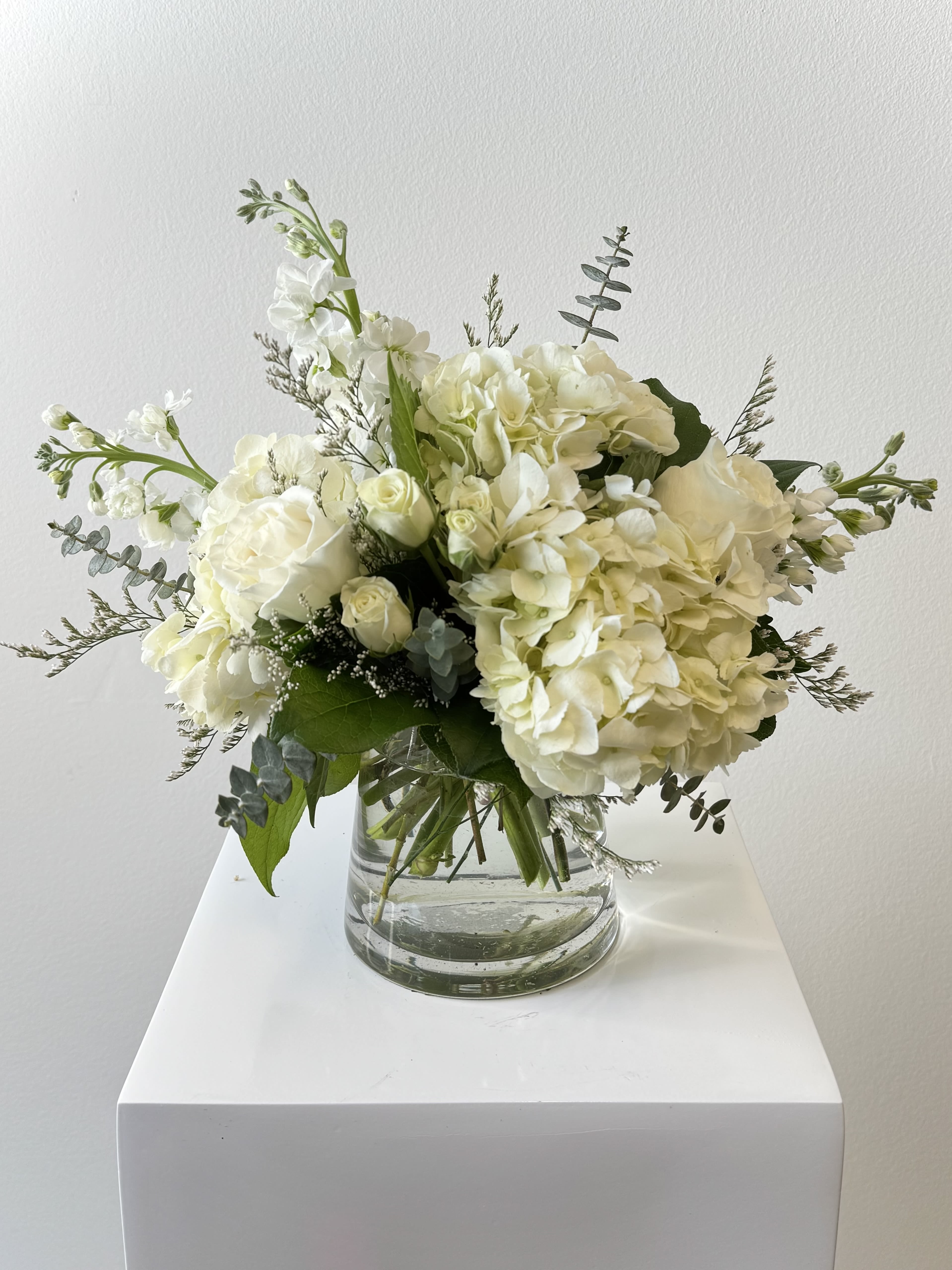 Simply Pure - A whisper of purity for gentle spirits. Soft and serene, the Simply Pure Bouquet from Irvine Village Flowers blooms with a delicate grace, blending pristine hydrangeas, tender stock, classic roses, and dainty spray roses, all kissed by subtle eucalyptus accents. This arrangement glows with a quiet elegance, a pure embrace that cradles the soul in moments of love and stillness.    The snowy hydrangeas unfold like soft clouds, symbolizing heartfelt gratitude, peace, and timeless devotion. The stock, with its gentle clusters, offers a breath of sweetness and calm, reflecting affection and harmony. Roses, pure and luminous, bloom with love and reverence, while spray roses sprinkle innocence and charm across the bouquet. Woven with the silvery touch of eucalyptus, they bring a soothing whisper of renewal and grace. Together, these blooms craft a vision of purity, a tender gift to uplift and cherish.    In its soft simplicity, this arrangement shines. Visit Irvine Village Flowers online or step into our store to embrace its tranquil beauty. With our gentle flower delivery across Irvine, your bouquet arrives fresh, a delicate treasure to share joy and light.