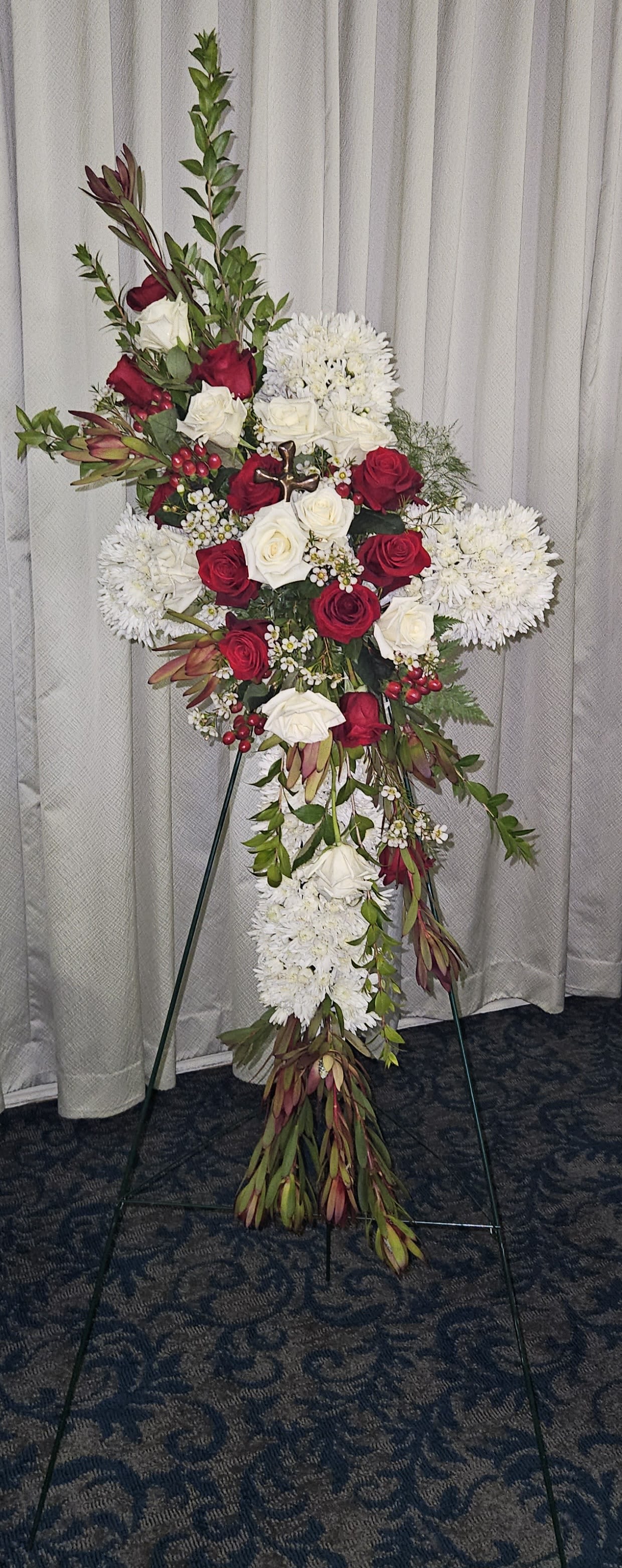 White and Red Cross Easel Spray - A grand, cross-shaped easel spray, created with white mums, red and white roses, and red hypericum berries. This spray is accented with a beautiful resin cross in the center, and is a perfect memorial for a loved one. 40in by a foot and a half.