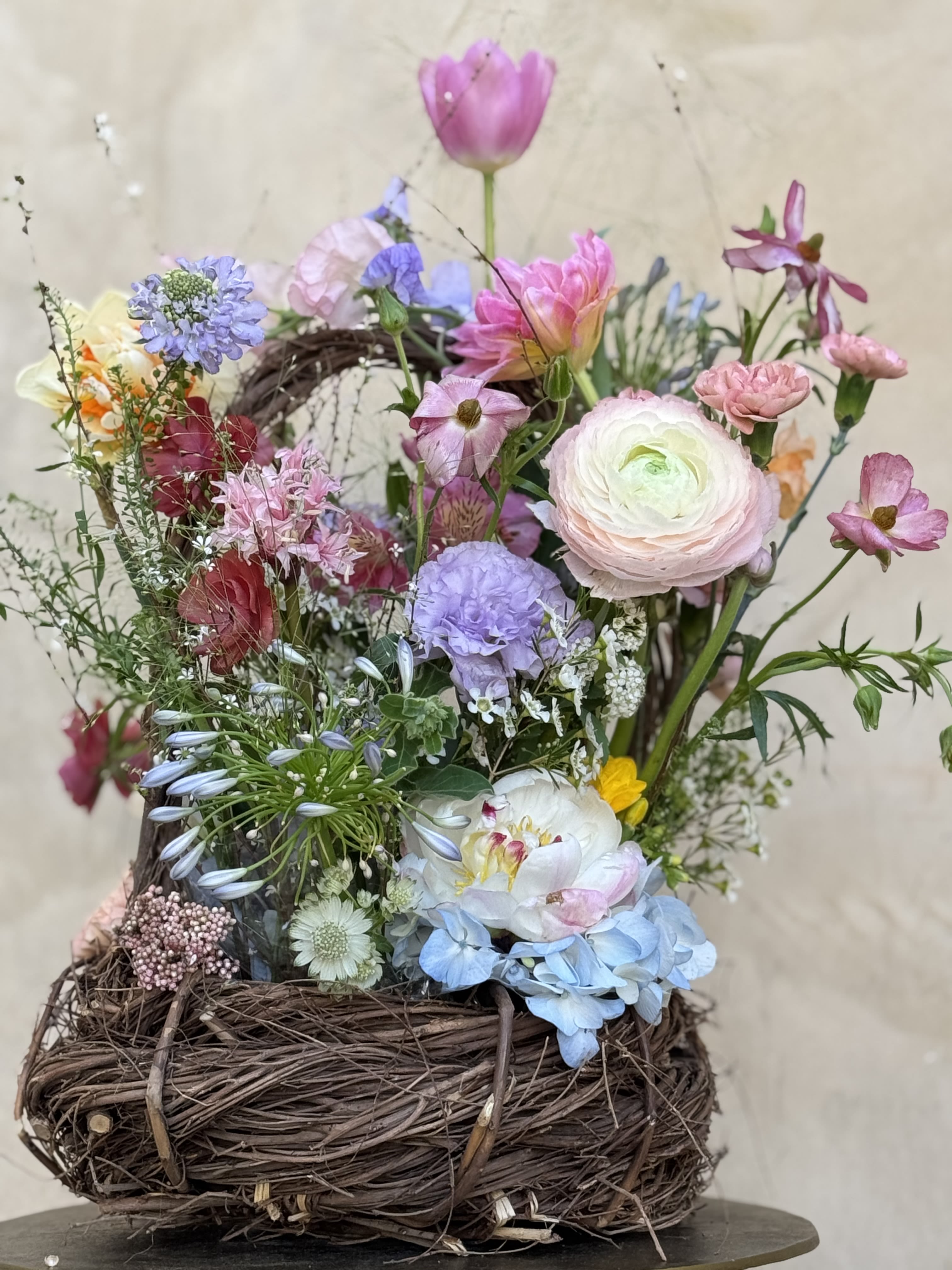 Touch of Nature - This charming rustic basket is filled with a vibrant mix of tulips ranunculus, and beautifully accented with delicate greenery. the assortment showcases an array of colors, including shades of pink, purple, yellow and cream, creating a whimsical and lively display. Perfect for adding a touch of natural elegance to any home or occasion
