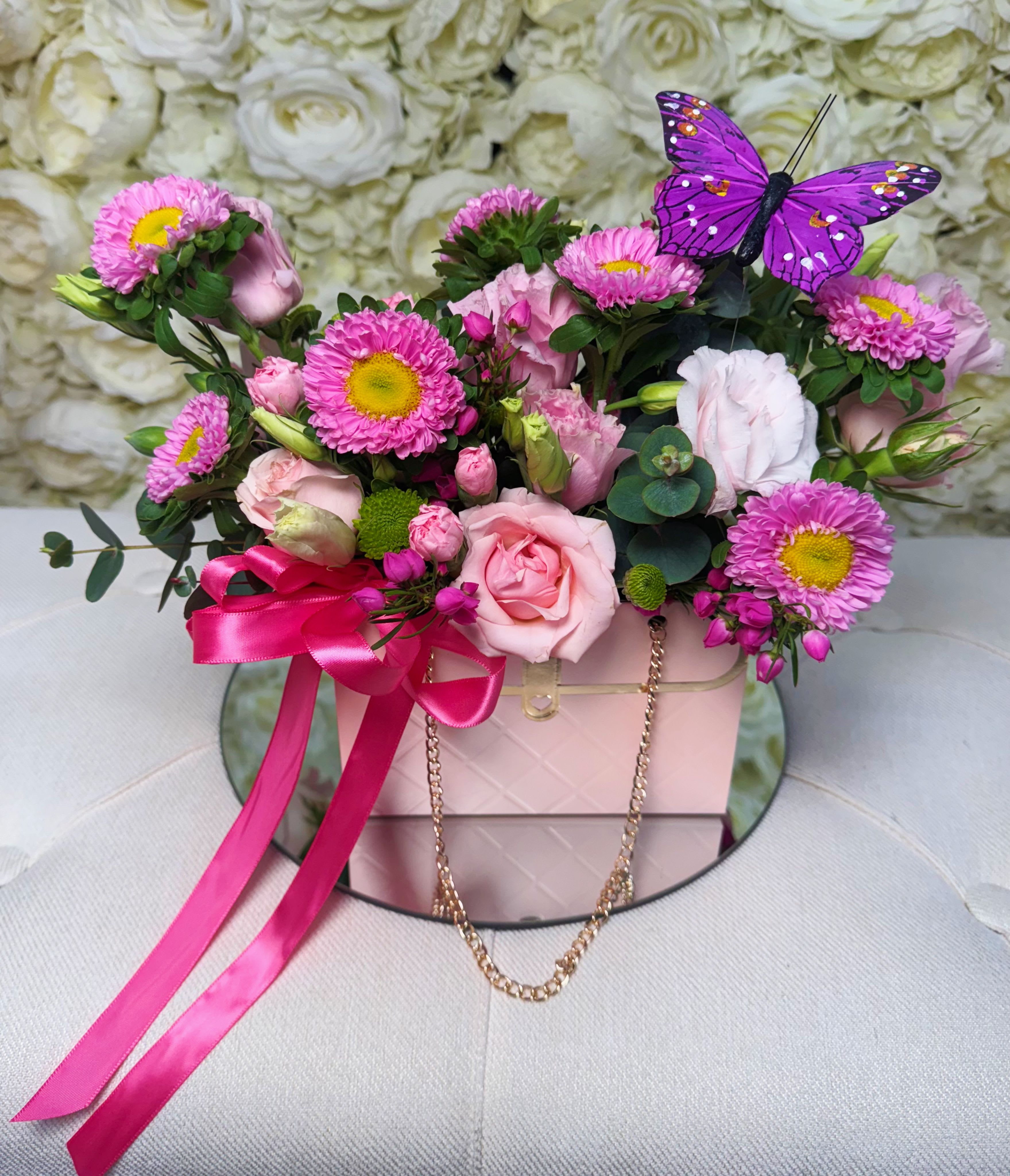 Sweet Charm - A charming and vibrant design, perfect for any special occasion. Crafted with spray roses, lisianthus, mini carnations, boronia, green button poms, and aster flowers, all beautifully arranged in a purse-shaped box with a gold chain, hot pink ribbon, and a butterfly for a magical touch. Ideal for gifting joy and sweetness.