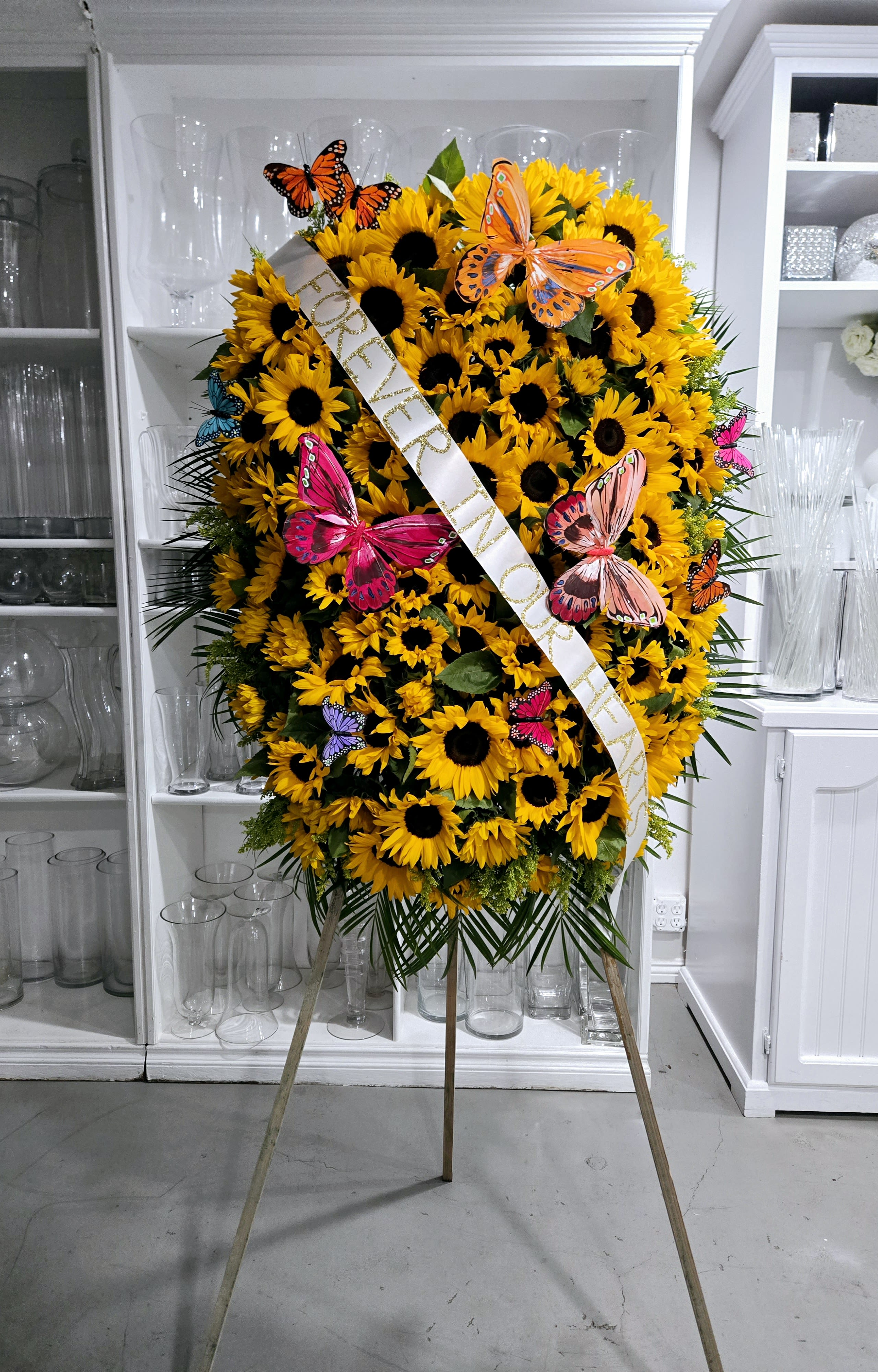 Sunlit Standing Spray - A heartfelt standing spray filled with bright sunflowers and adorned with colorful butterflies. A symbol of warmth, joy, and remembrance—perfect for celebrating a life that brought light to others.  Standing Spray