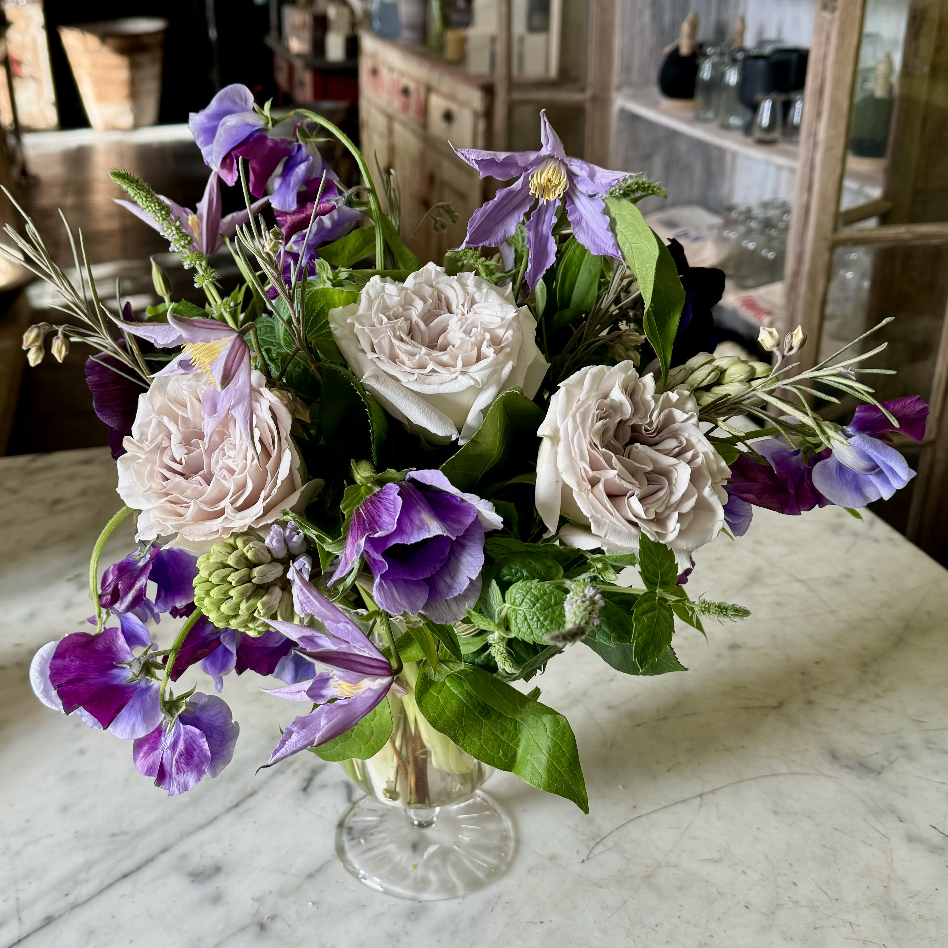 Urban Gardener Lavender Haze - A footed delicate glass vase filled with seasonal blooms in lavenders to purples. Daily flowers may vary!