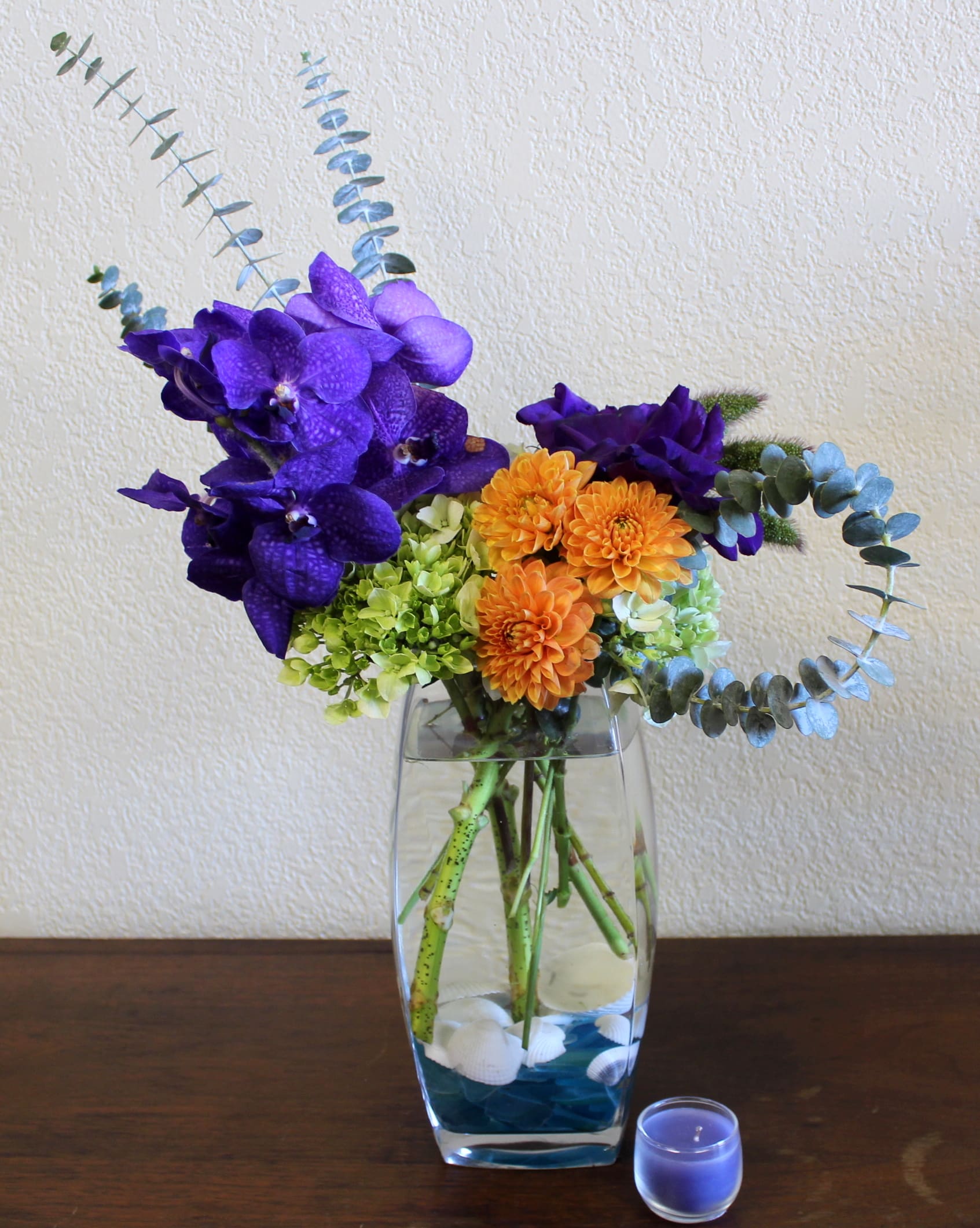 Oceanic Bliss Oceanic Bliss - Exotic orchids on a bed of hydrangea. pops of orange accented by eucalyptus. seashells and blue sea glass make this arrangement complete!