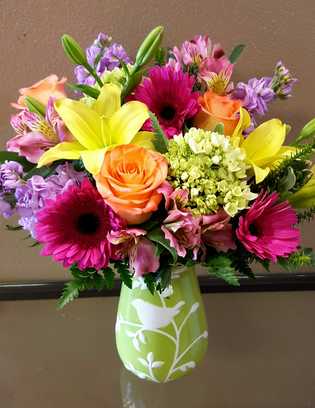 SPRING PERCH - A LOVELY GREEN VASE, WITH A LUSH MIX OF LILIES, GERBERAS, ROSES, STOCK, AND OTHER SEASONAL FLOWERS.  